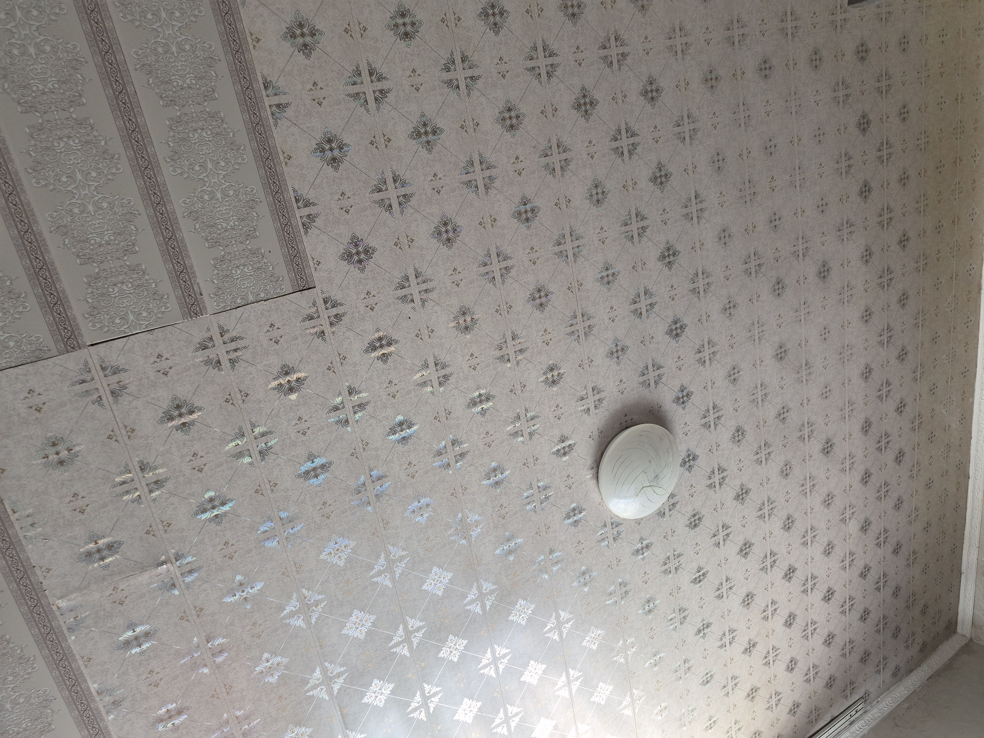 This media file shows an indoor ceiling with patterned wallpaper and a light fixture. The wallpaper has a repeating geometric design, with some sections featuring a more intricate, ornamental pattern. The colors are muted, with shades of grey and beige dominating. A circular, white light fixture with a ribbed texture is attached to the ceiling. The overall impression is of a typical interior space, possibly a room in a residential or commercial building. There is no indication of the time of day or weather outside. The location is identified as Қанабек, Kazakhstan. There is no visible text in the image.