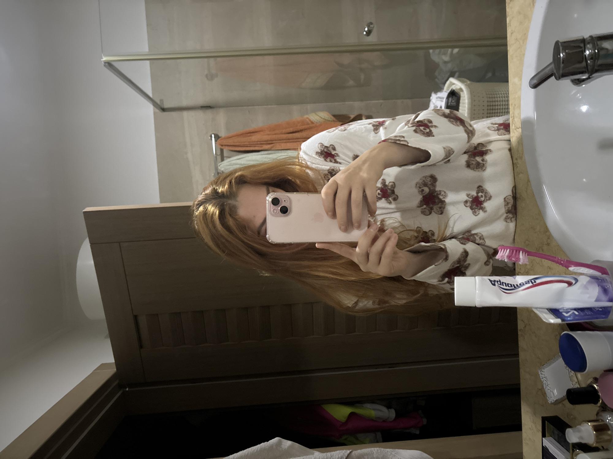 A red-haired female takes a selfie in a bathroom in Budva, Montenegro. The camera is a pink iPhone with visible lenses. She is wearing a teddy bear-printed robe. The bathroom has a sink, a cabinet with clothes inside, a toothbrush, and toothpaste. A towel rack with towels is in the background behind a glass partition.