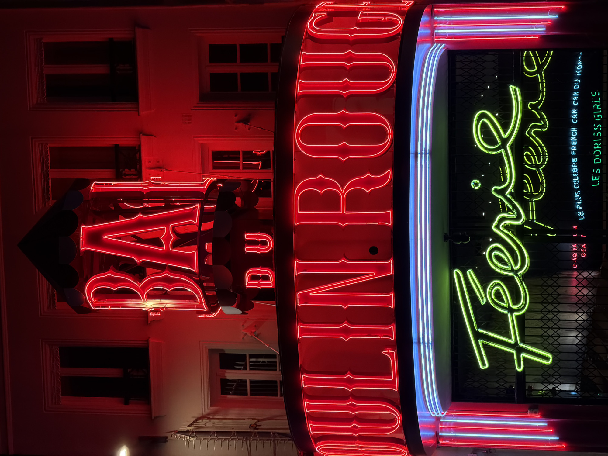 A nighttime scene showcases the iconic neon signage of the Moulin Rouge cabaret in Paris, France. The dominant feature is a large, vertical red neon sign spelling "MOULIN ROUGE," illuminating the white building facade with a deep red glow. Adjacent to it, a vibrant green neon sign in stylized cursive script reads "Féerie," which is the title of the cabaret's show. Below and to the right of "Féerie," smaller neon text in white states "LE PLUS CELEBRE FRENCH CAN CAN DU MONDE" (The Most Famous French Can Can in the World), and in green, "LES DORISS GIRLS," referring to the venue's dancers.
Partially visible on the left, another red neon sign displays "BAL DU," likely part of a phrase like "BAL DU MOULIN ROUGE." The signs are set against a dark background, indicating it is night, with glimpses of closed windows and building architecture. Blue and white neon accents run along the edges of some signs, adding further dimension to the display. No people or specific activities are visible, but the bright, colorful neon lights vividly advertise the world-renowned entertainment venue, evoking the lively and historic Parisian nightlife.