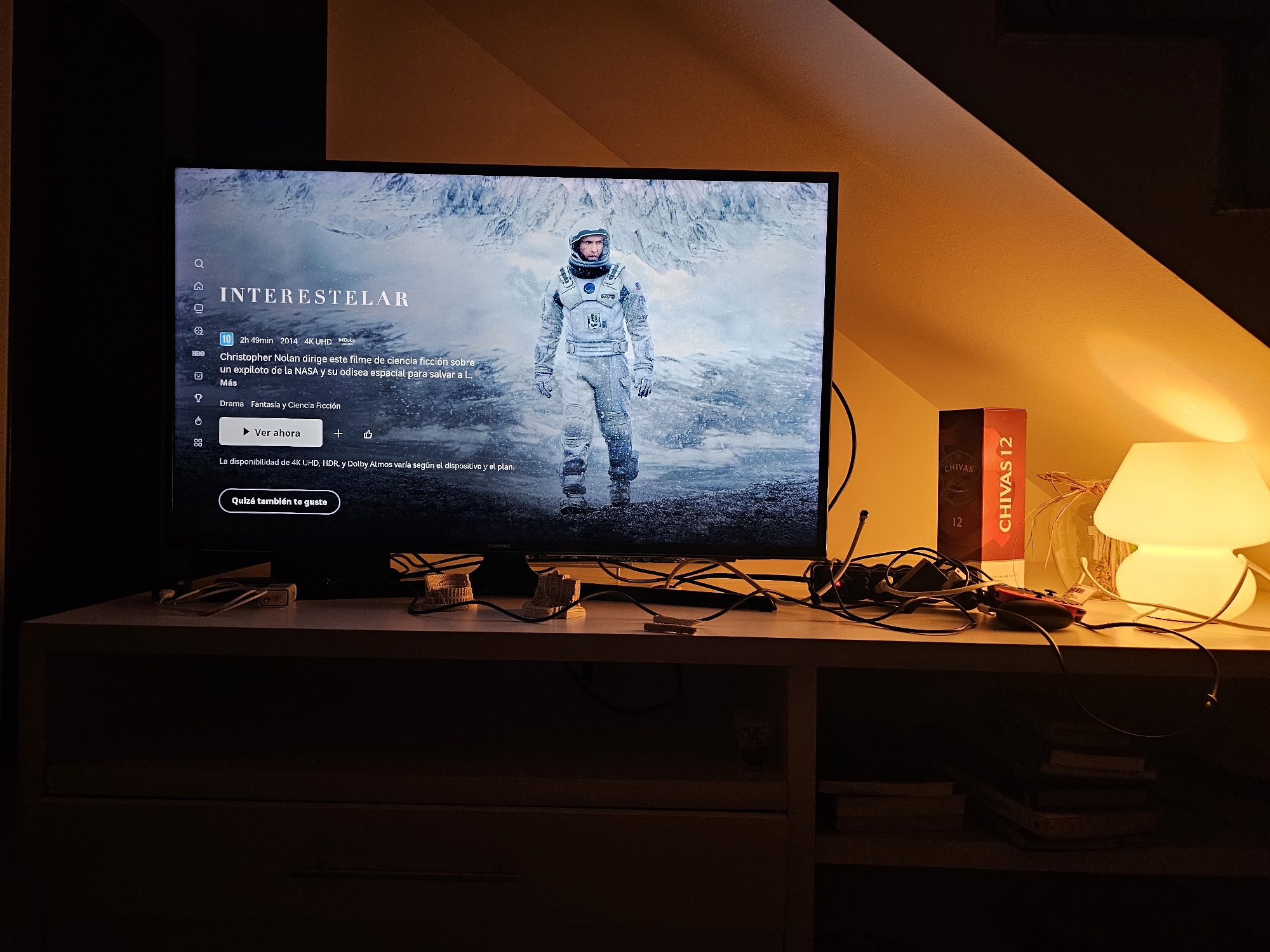 A dimly lit interior shot shows a television displaying the title screen for the movie "Interstellar," along with a description in Spanish. The screen is framed by a black bezel. The movie's image shows a person in a spacesuit standing in a snowy, icy environment. The television is placed on a light-colored shelf with a drawer visible at the bottom. Wires and electronics are scattered on the shelf in front of the television. To the right of the television, a box of Chivas Regal 12 Scotch Whisky is visible, along with a small white lamp emitting a warm glow. Below the shelf, books are stacked. The lighting is warm, casting shadows that suggest an evening or nighttime setting.