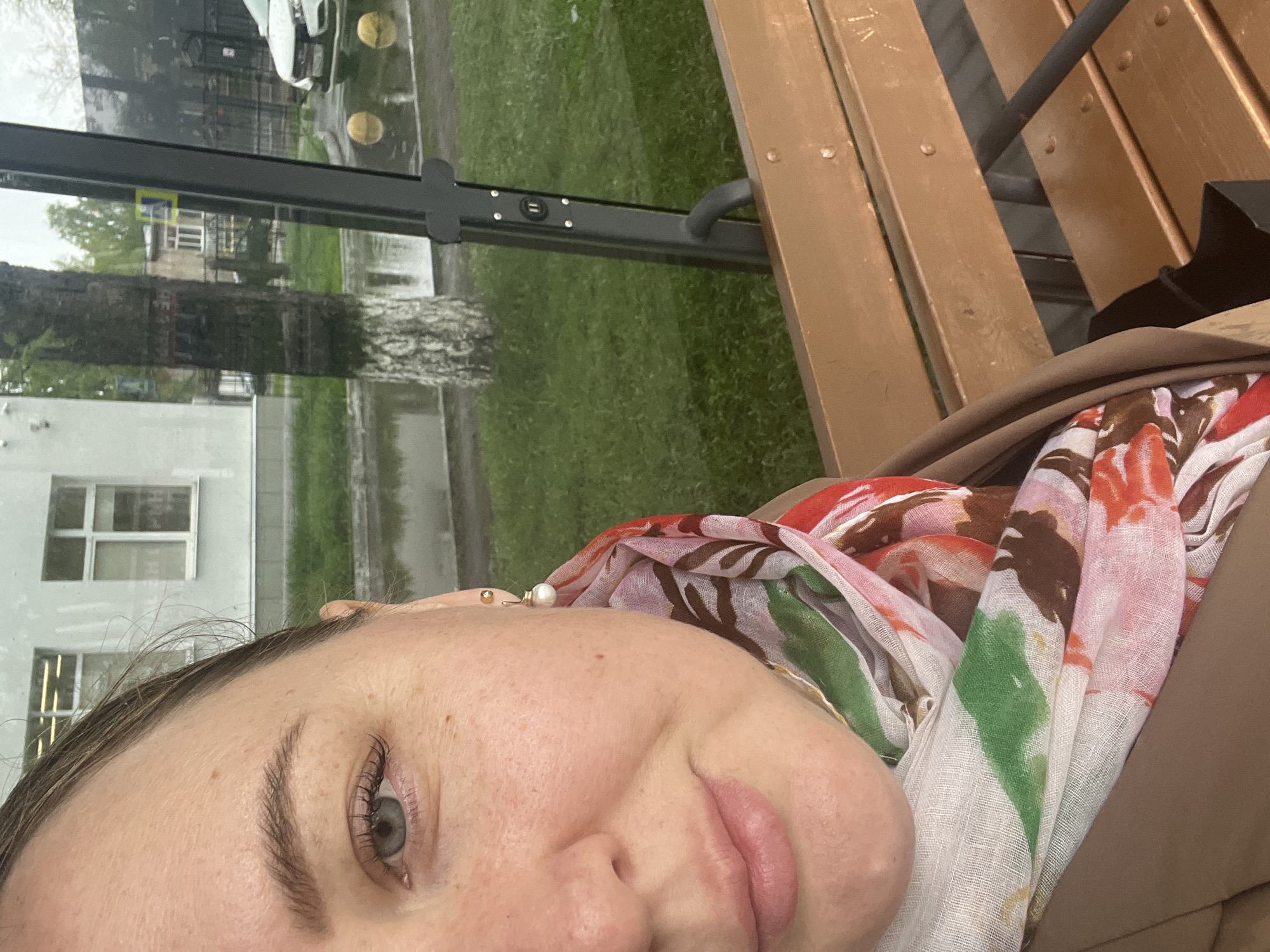 A person is sitting on a park bench next to a bus shelter in Chaikovsky, Russia. The view through the shelter's glass shows a birch tree and a building. The person is wearing a scarf with floral patterns. The bench is made of wood. The weather seems overcast.