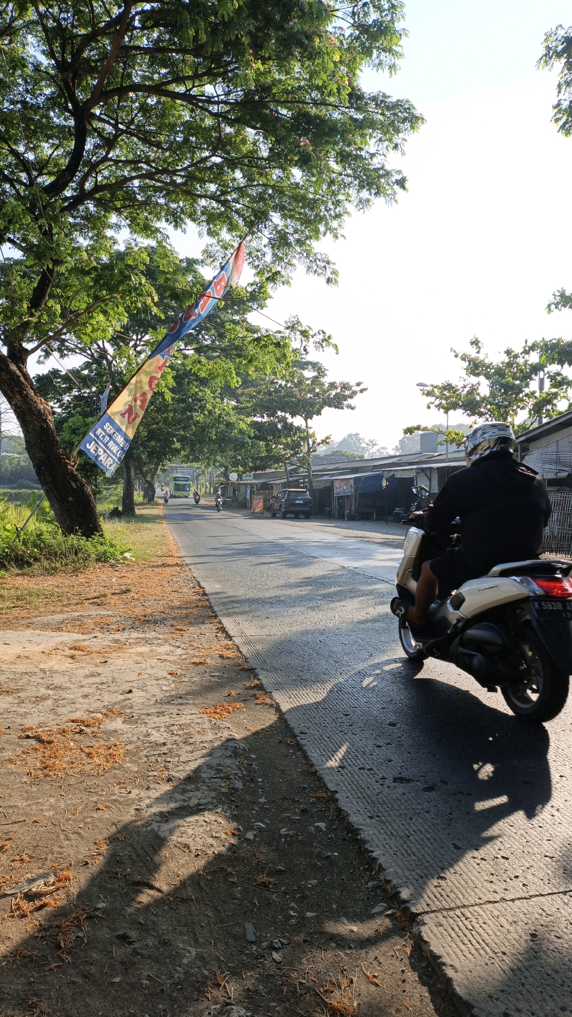 A motorcycle rider wearing a black jacket and helmet rides a white motorcycle along a paved road in Bandengan, Indonesia. The road is lined with trees on the left side, with a banner hanging from one of them. On the right side of the road are several small buildings that appear to be shops. The sun is shining, casting long shadows on the road and the ground. There are a few other vehicles and riders visible in the distance, and the sky is bright and clear. The ground on the left side of the road is dirt and covered with fallen leaves.