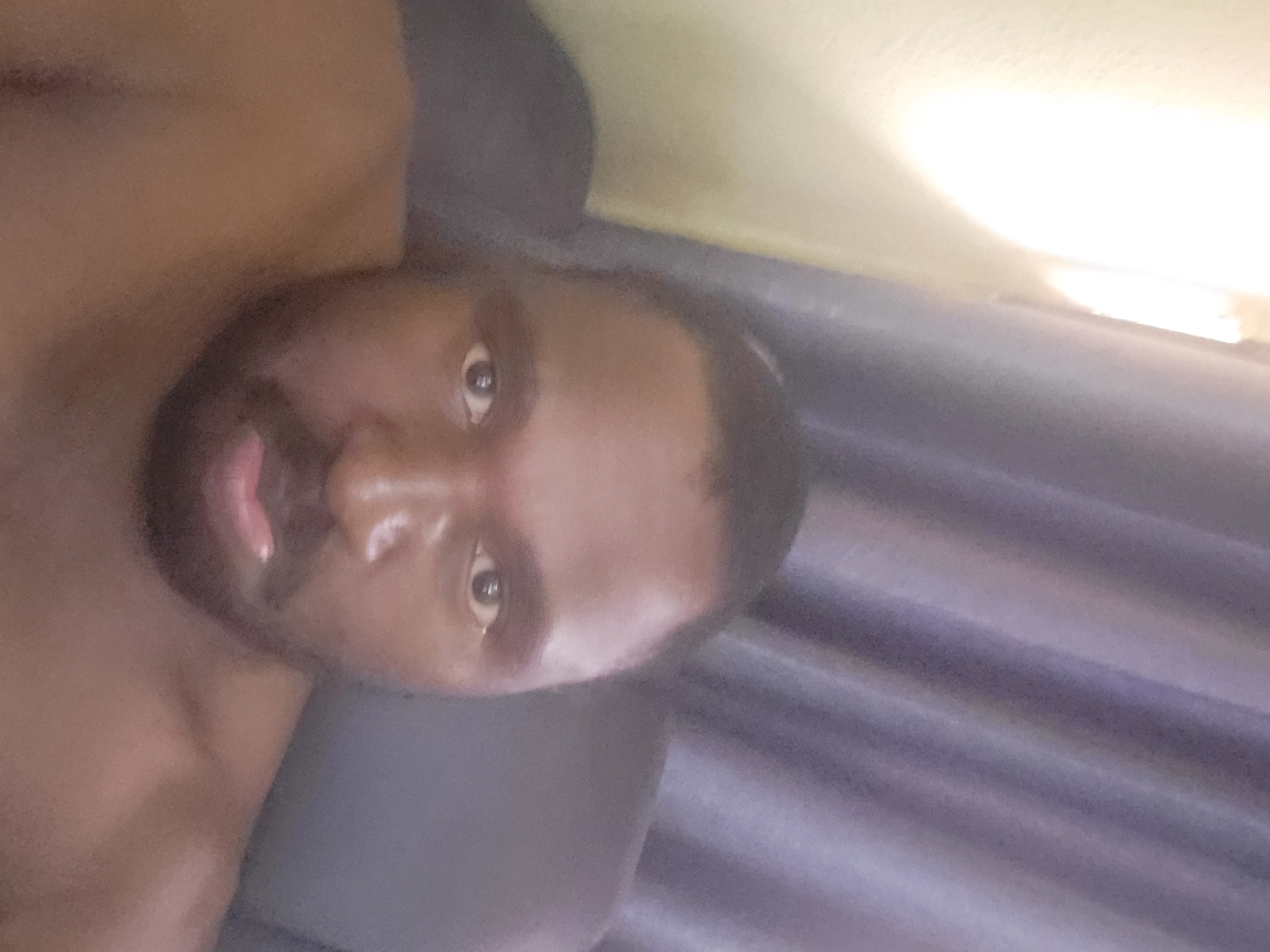 This is a close-up, low-angle, portrait-oriented media file of a Black man. The man's head is tilted slightly, and he appears to be looking directly at the camera with a neutral expression. He has a beard and short hair. His skin is brown, and his eyes are dark. The lighting in the image is strong, creating a highlight on his forehead and nose.
The background is out of focus but appears to consist of dark curtains or fabric on the right side of the frame and a lighter, possibly yellowish surface on the top. The scene is indoors, and given the lighting, it could be daytime or a well-lit room at night. There is no visible text in the media file. The general context suggests a casual or personal setting, possibly a home environment in Suleja, Nigeria. The file is rotated 90 degrees clockwise, with the man's head to the left.