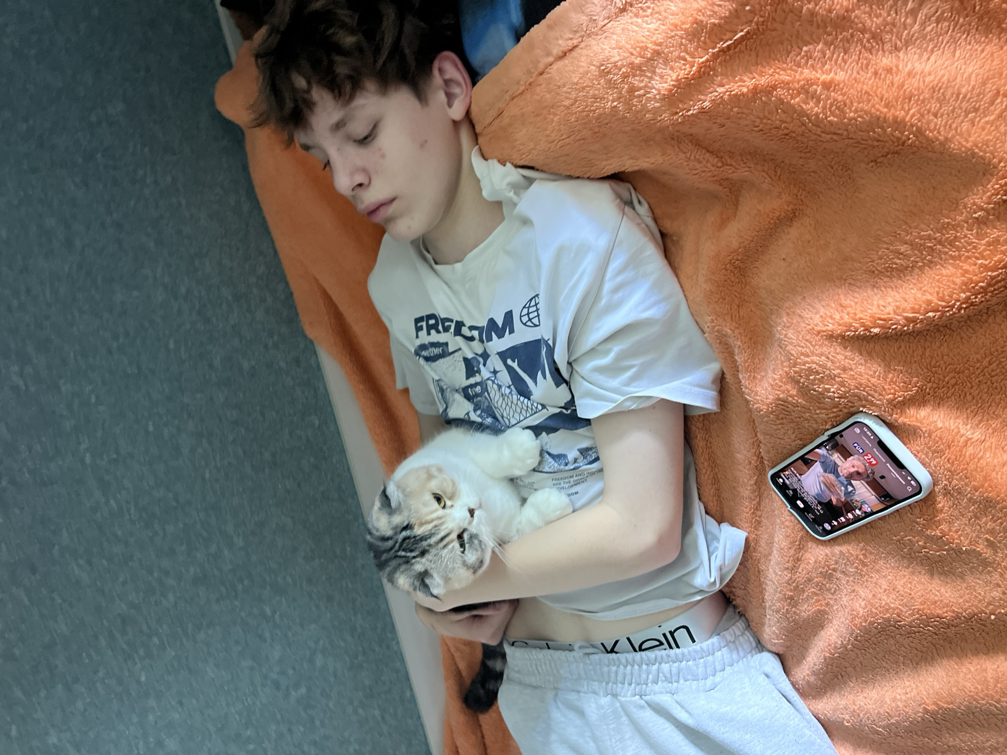 Here is a detailed description of the image:
**Scene:**
The image depicts a person lying down with a cat and a smartphone on an orange blanket. The scene appears to be indoors, possibly in a bedroom, as there is a wall visible on the left side of the frame.
**People:**
There is one person visible, a young individual with short, curly hair who appears to be sleeping. The person is lying on their back, wearing a white t-shirt with the word "FREEDOM" printed on it, along with grey shorts. The brand "Calvin Klein" is visible on the waistband of the shorts.
**Subjects:**
A cat is nestled in the person's arms. The cat is white and grey.
**Objects:**
A smartphone lies on the blanket next to the person. The phone's screen is visible, showing a video with the text "FÜR 2.99". The blanket has a soft, plush texture.
**Actions:**
The person is sleeping. The cat appears to be resting comfortably in the person's arms.
**Interactions:**
The person and cat are shown in close proximity and appear to be sleeping together.
**Environment:**
The environment appears to be a bedroom, although specific details are limited. The wall on the left has a bluish-grey pattern.
**Location:**
No specific landmarks or visual elements pinpoint the location to Noviy, Russia.
