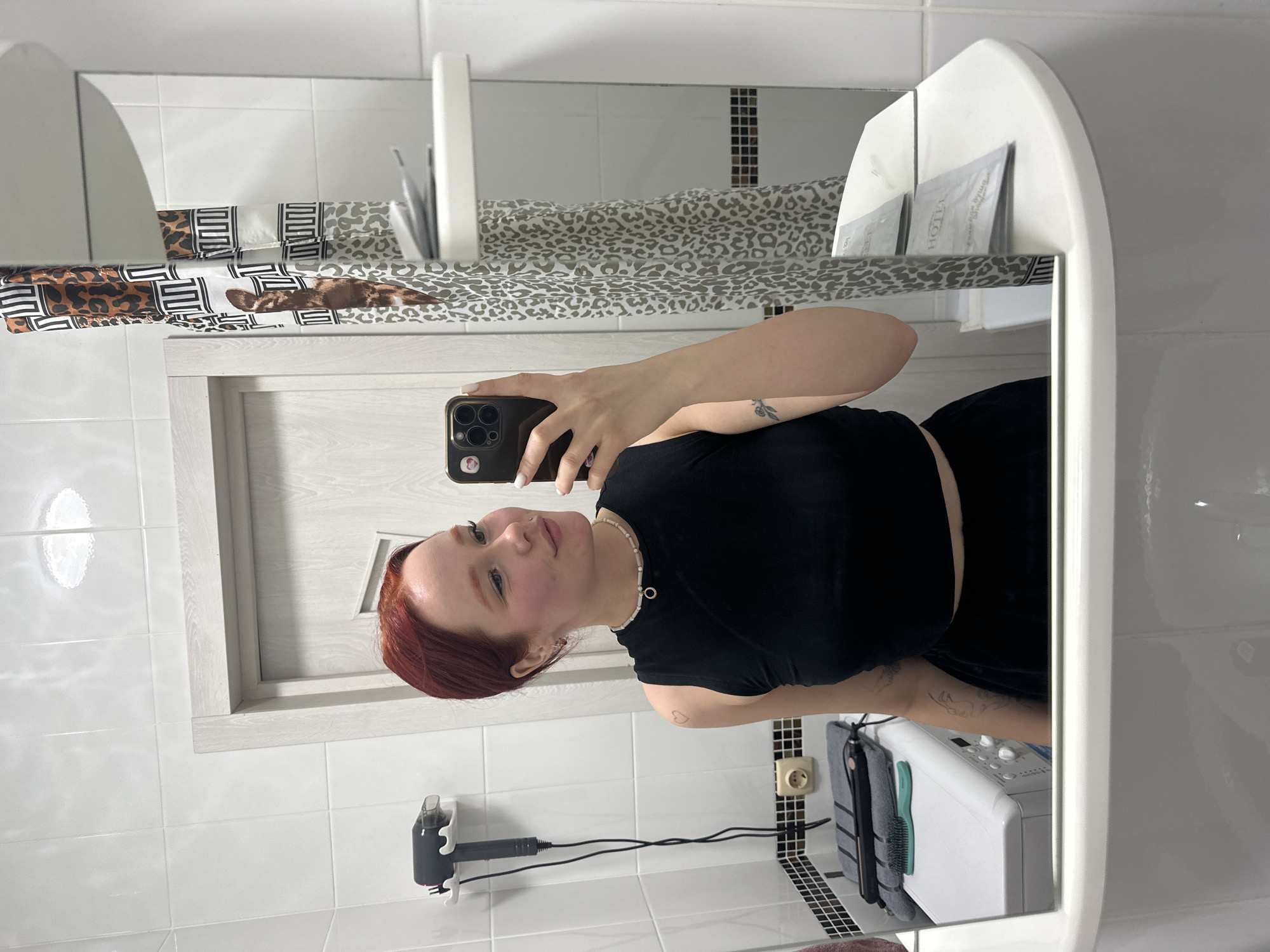 A young Caucasian woman with short, red hair is taking a mirror selfie in a bathroom. She is wearing a black crop top and a black skirt. The bathroom has white tiled walls and a patterned shower curtain. In the background, a washing machine and a hairdryer are visible. The woman has a necklace with a circular pendant. She is smiling slightly and looking at her reflection.