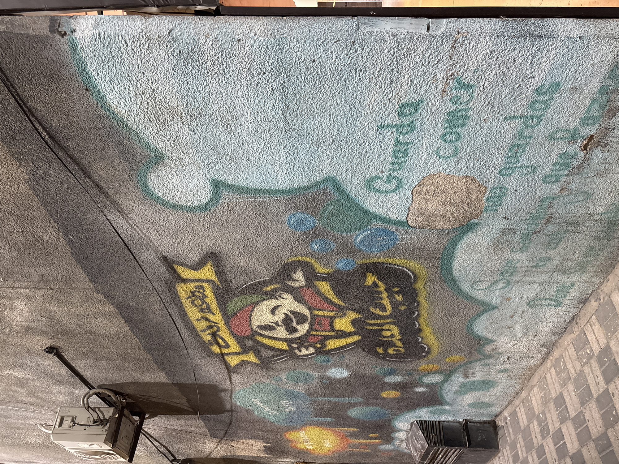 The image depicts a textured, grey concrete wall with graffiti art. On the left side, an outdoor air conditioning unit is mounted on a black bracket. The wall features a yellow banner with Arabic script, followed by a cartoon character with a mustache wearing a red and yellow hat. Below the character, more Arabic script is visible. To the right of the character, there are spray-painted blue and green bubble-like shapes. Further to the right, the wall is painted a light blue-grey, with the words "Guarda Comer" and some faint Arabic script in a teal color. The ground in front of the wall is paved with rectangular grey and beige bricks. The scene is lit by artificial light, casting shadows on the wall. This graffiti is located in Amman, Jordan.