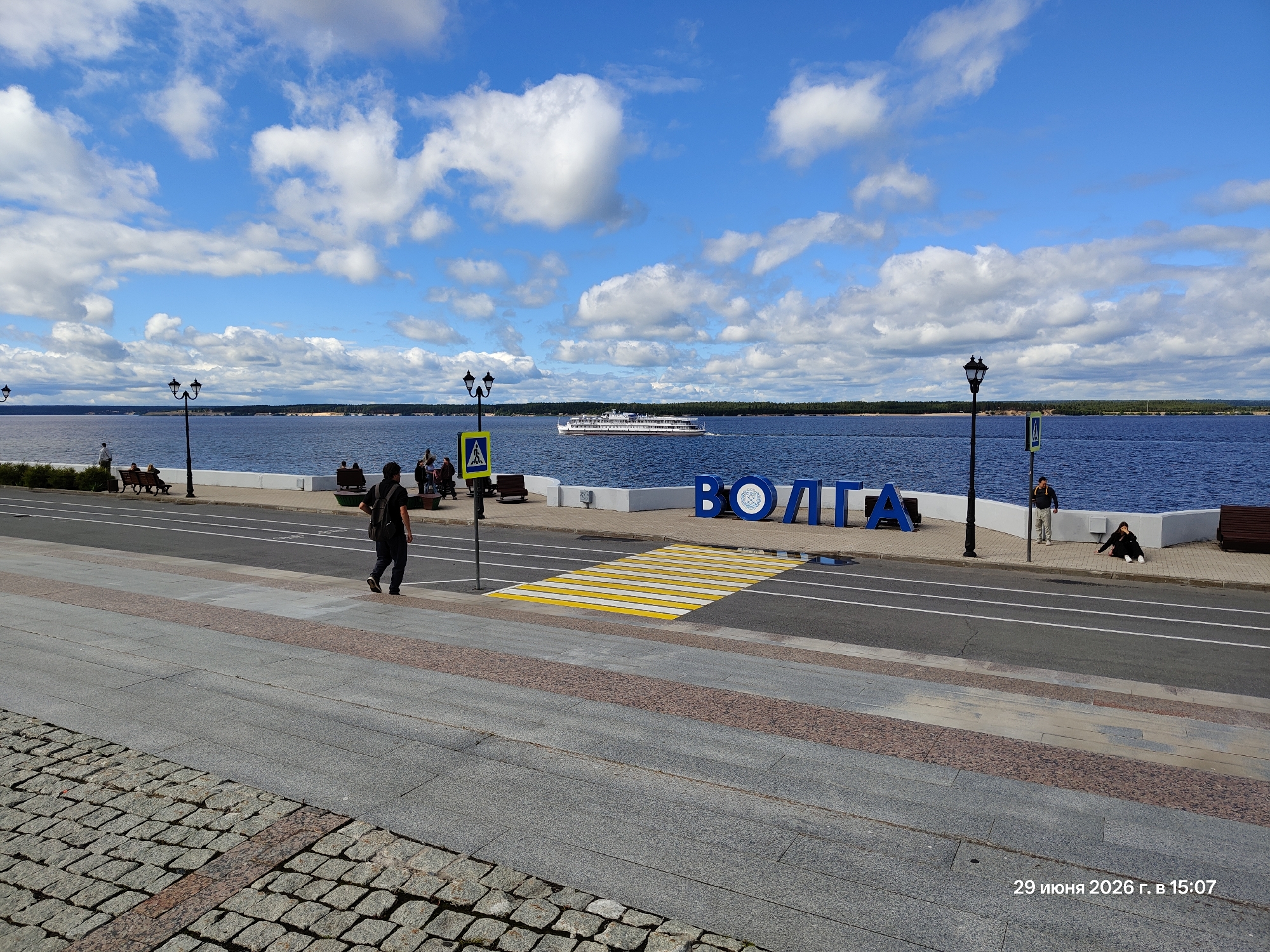 The image depicts a waterfront scene on a sunny day with scattered clouds. The foreground is paved with cobblestones, leading to a gray paved walkway bordered by a reddish stripe. Beyond the walkway is a road with white lane markings. A pedestrian crossing with bright yellow and white stripes is prominently featured on the road.
In the middle ground, a wide blue body of water, likely a river or lake, stretches to the horizon. A large white passenger boat with multiple decks is sailing on the water. Along the waterfront, there is a white embankment with lampposts. Large blue letters spelling out "ВОЛГА" (Volga) are visible on the embankment, serving as a prominent landmark.
Several people are present in the scene. On the left side, a man with a backpack is walking on the walkway. Further back, some people are sitting on benches. On the right side of the embankment, a man is standing, and a woman is seated on the ground.
The sky is a vibrant blue with fluffy white clouds, suggesting pleasant weather. The time of day appears to be afternoon, as indicated by the bright sunlight and shadows. The overall mood is peaceful and leisurely, typical of a summer day at a scenic waterfront. The visible text "29 июня 2026 г. в 15:07" indicates the date and time the photo was taken.