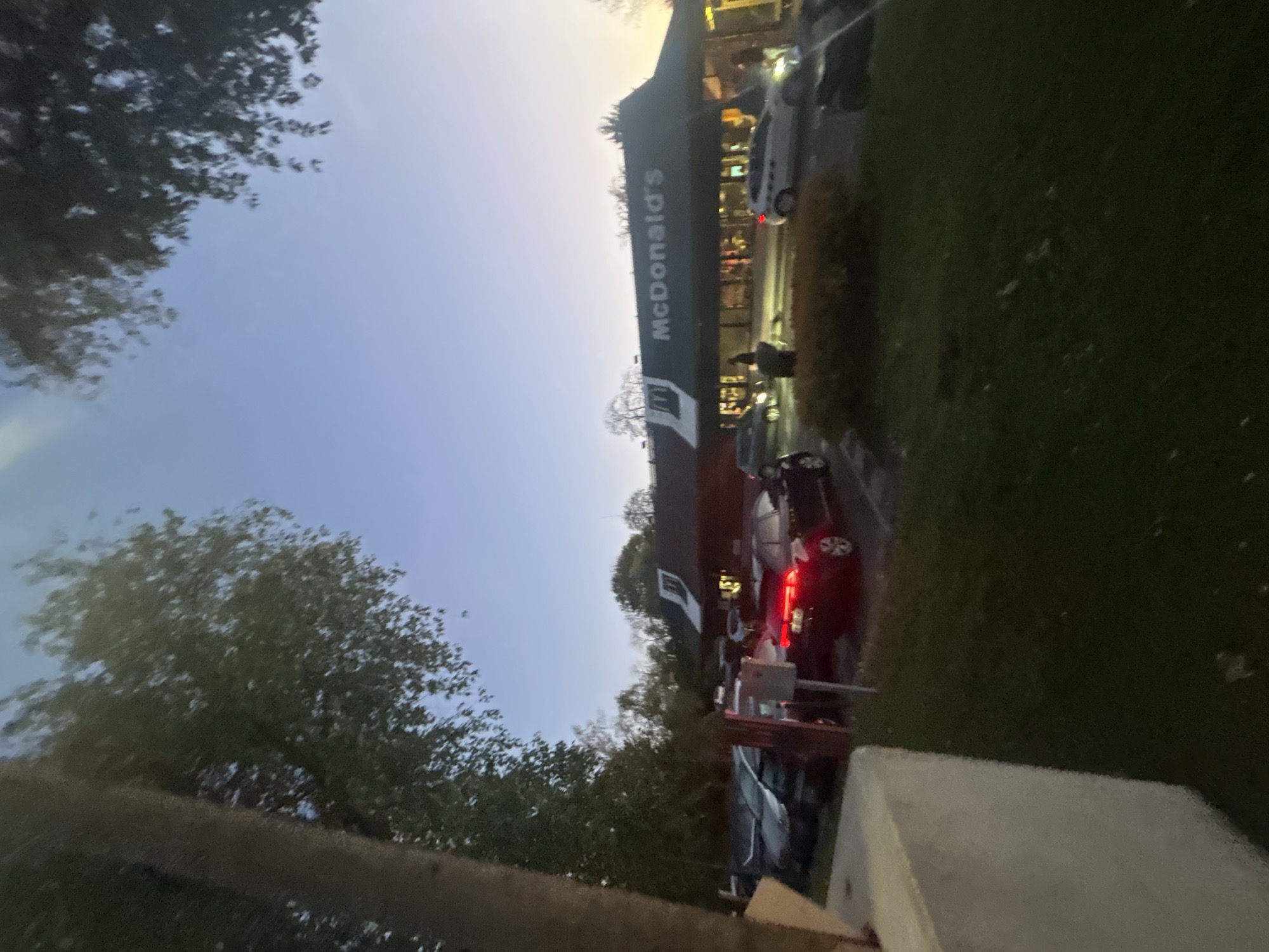 The image depicts a McDonald's restaurant, likely located in Noisy-le-Grand, France, given the presence of cars with European license plates. The building is situated on a street with parked cars in front of it. A line of cars is in the drive-through. The sky is dusky, suggesting the time is either early morning or late evening. Trees are visible on both sides of the street.