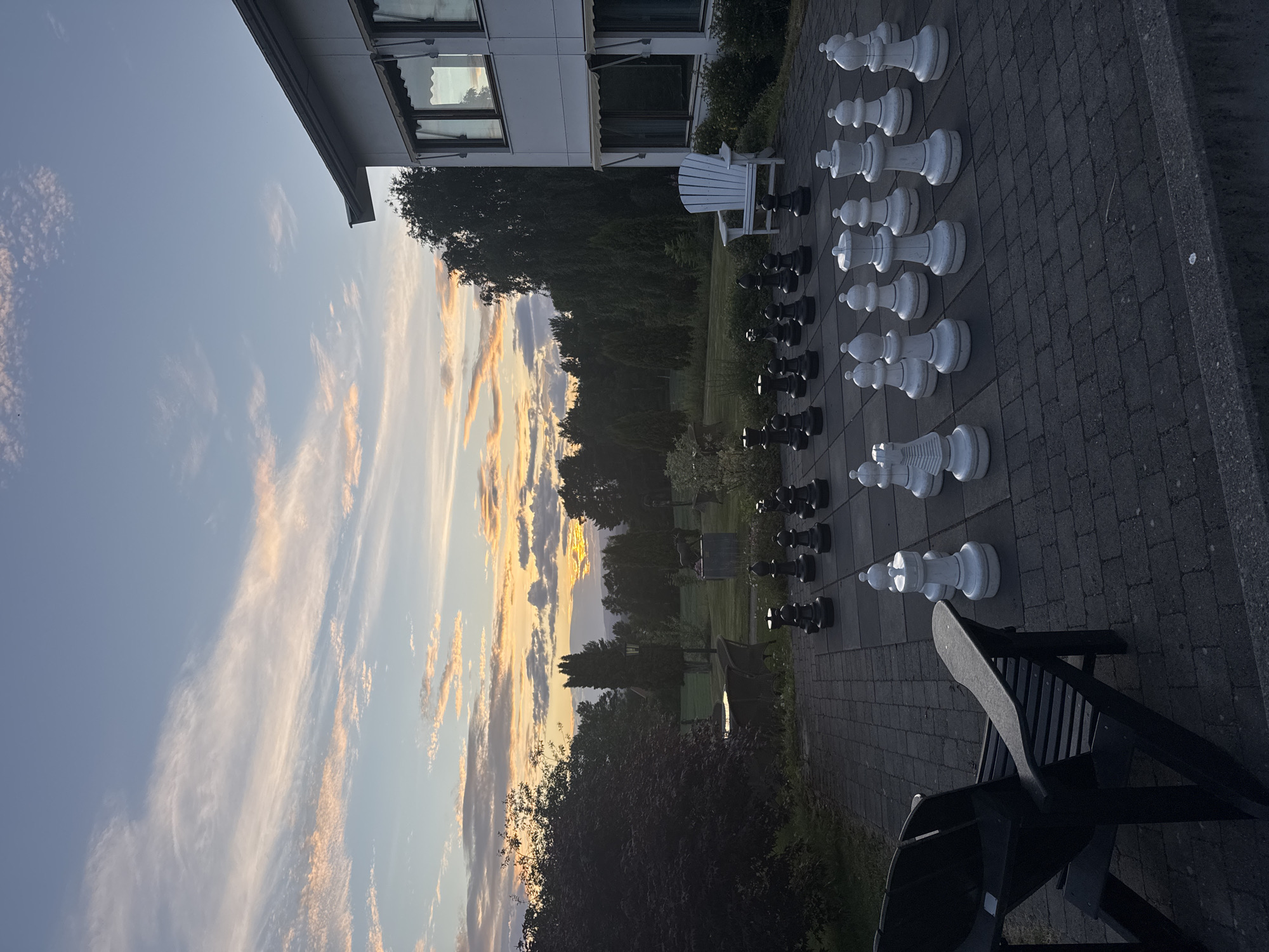 The image depicts an outdoor oversized chess set on a paved patio in front of a modern white building. The chess pieces, white on one side and black on the other, are arranged on the board, suggesting a game in progress or recently played. A white Adirondack chair is placed near the chess board, and a dark Adirondack chair is visible in the foreground. The background features a landscape with trees and a distant view of hills under a colorful sunset sky with clouds. The overall scene suggests a serene evening setting.
While the image captures a visually appealing scene, it does not contain any specific elements that would allow for the definitive identification of its location within Hønefoss, Norway. The architectural style of the building, the types of trees, and the general landscape are common features found in many locations and do not provide unique identifiers for Hønefoss. Therefore, it is not possible to confirm this specific location.