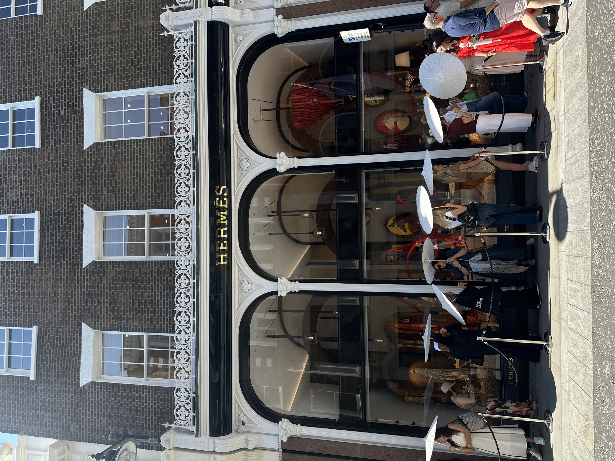 The image shows the exterior of the Hermès store on Bond Street in London, United Kingdom. The store features a dark brick facade with large arched windows. Above the windows, the word "HERMÈS" is displayed in gold lettering on a black background. A decorative white railing runs along the top of the store front.
In front of the store, a line of people are waiting, many holding white parasols to shield themselves from the sun. The people are dressed in various casual and formal attire. The pavement in front of the store is paved with light-colored stones, and stanchions with ropes delineate the queue. The environment appears to be a sunny day.