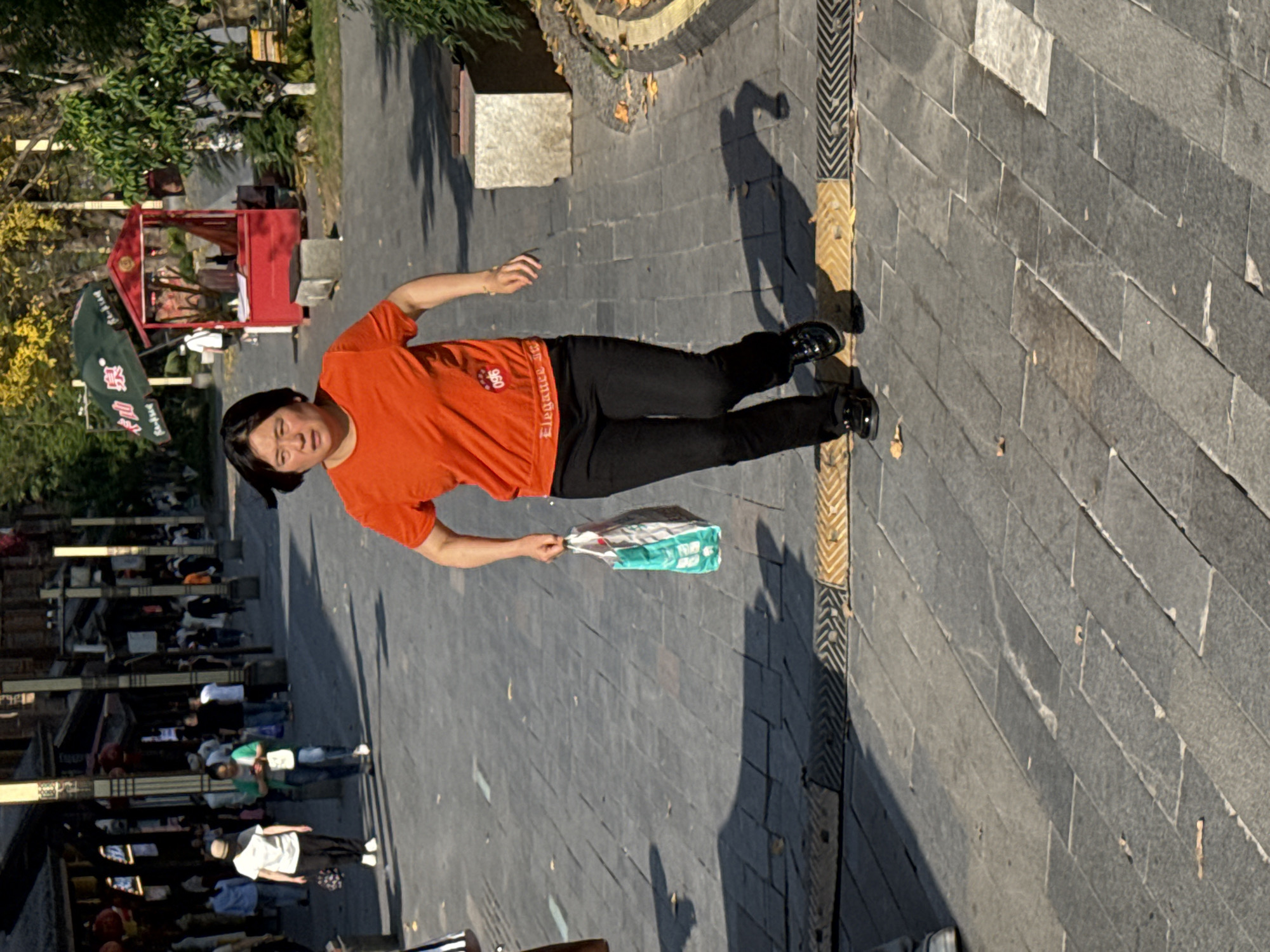 An outdoor scene in Shapingba District, China, depicts a bustling public area under bright sunlight. The image is oriented vertically (rotated 90 degrees clockwise).
The main subject is an individual with short dark hair, wearing an orange t-shirt and black pants, walking forward. They hold a white and turquoise plastic bag in their left hand. The orange t-shirt bears partially visible white text, likely "Elegance," and a "960" logo. Their expression is neutral. Long, distinct shadows are cast on the paved ground, indicating a strong light source from the side, suggesting it is midday or afternoon.
The setting is a pedestrian area paved with rectangular grey and lighter-toned tiles. Along one edge of the pavement, there's a distinctive yellow and black patterned strip. In the background, numerous other people are visible, some standing, many seated at tables or low structures, suggesting a market, park, or outdoor dining area. Further in the background on the left, a small, distinct red kiosk with a pointed roof and a green banner featuring Chinese characters is visible amidst trees and foliage, some of which show autumnal yellow leaves.
The overall atmosphere is that of a lively public space, with individuals going about their daily activities. The weather appears clear and sunny.