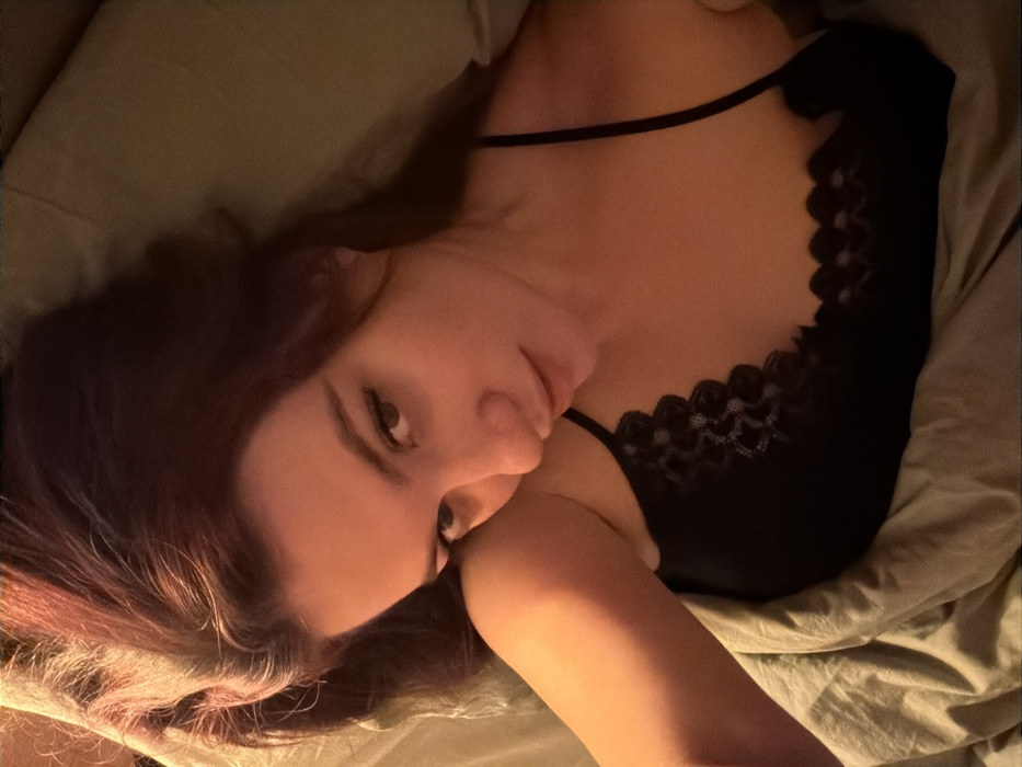 A woman with auburn hair lies on her side on a bed with green sheets, gazing at the camera. She wears a black lace-trimmed camisole. Her head is tilted, and her left hand is near her face. The lighting is subdued, creating soft shadows.