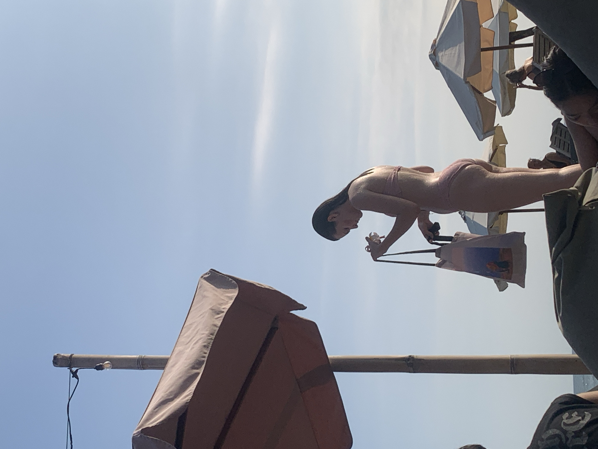 In a bright, sunny scene at North Kuta, Indonesia, a person is captured mid-action, seemingly performing a dive or a similar acrobatic maneuver against a clear blue sky. They are wearing a light-colored bikini and their body is arched backward, indicating a dynamic pose. Their long, dark hair streams behind them, adding to the sense of movement.
The person is holding onto the straps of a patterned tote bag, which dangles below them. The bag appears to have an image on it, possibly a landscape or artwork, but it is not clearly discernible. In the background, several large beach umbrellas with beige and white canopies are visible, suggesting a resort or beach setting. Some of these umbrellas are tilted, hinting at a slightly breezy day.
Partially visible in the foreground and to the sides are structures that appear to be part of the beach umbrella framework, possibly made of bamboo or similar material. A single light bulb is also visible attached to a horizontal bamboo pole. Other individuals are faintly visible in the background, some seated under umbrellas, suggesting a public recreational area. The overall impression is one of a leisurely day at the beach, with an emphasis on water sports or recreational activities, possibly a diving exhibition or a training session. The bright sunlight and clear sky indicate it is daytime, likely mid-morning or afternoon.