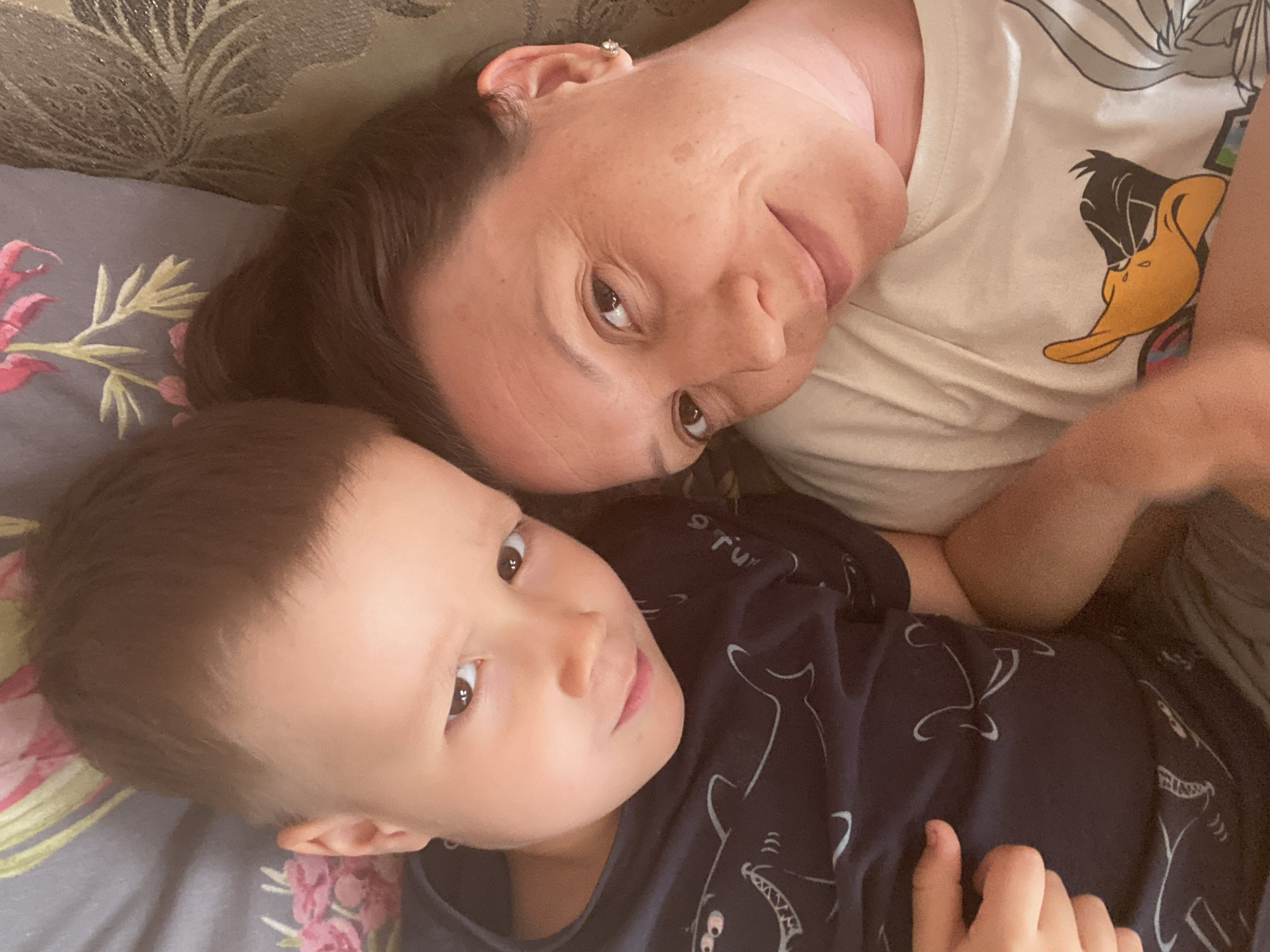 The image depicts a woman and a young child lying down together, likely on a couch or bed, with the woman's face positioned at the top of the frame and the child below. The woman is wearing a white t-shirt with a cartoon character (possibly Daffy Duck) and an earring. The child is wearing a dark t-shirt with white shark illustrations. The background includes a floral-patterned pillow and a fabric with a leaf pattern. The location in Noviy, Russia cannot be inferred from the image.