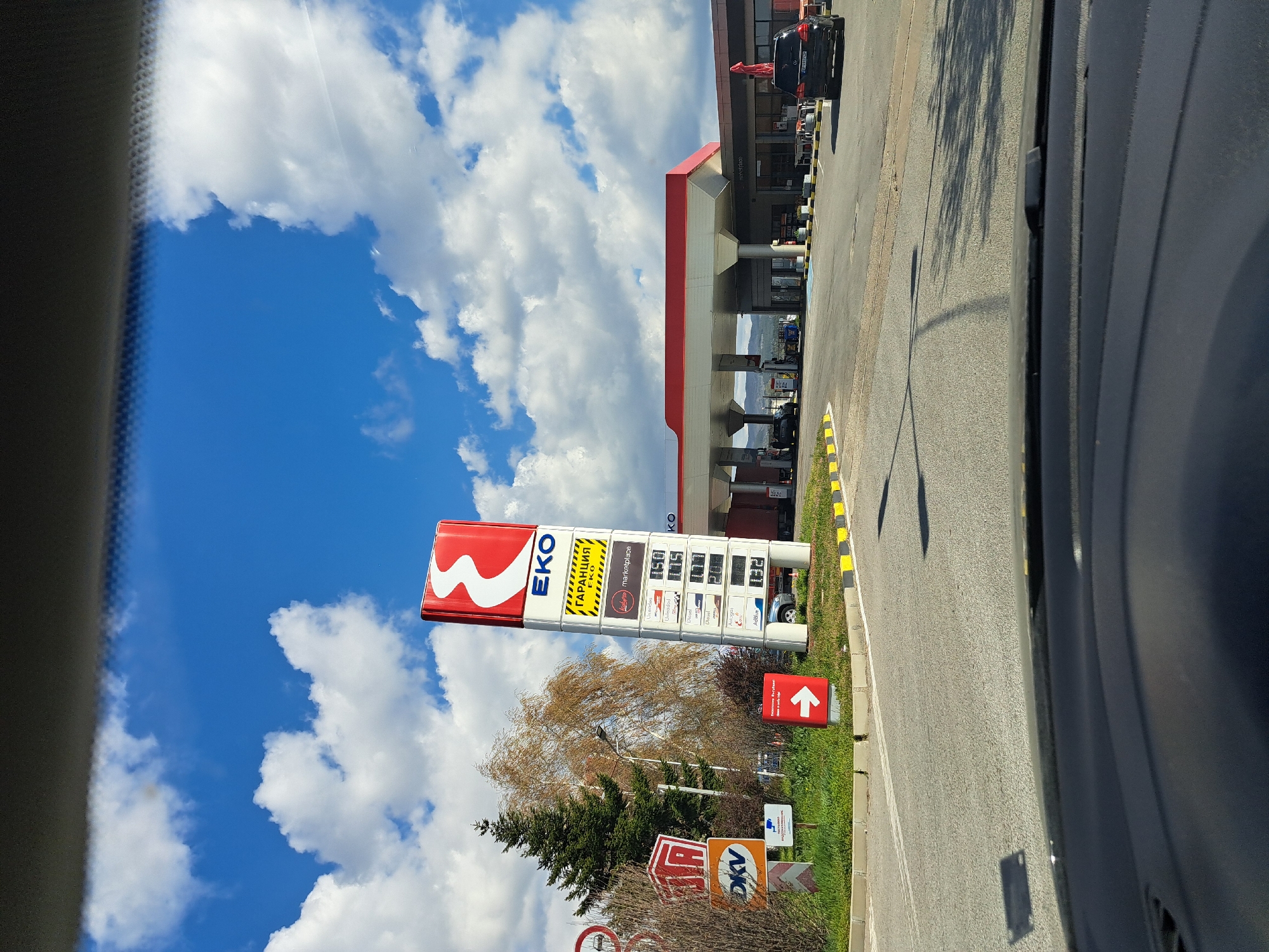 The image shows a roadside scene at an EKO gas station. The sky is blue with scattered clouds. A large EKO sign displaying fuel prices is visible, alongside other road signs. Cars are parked at the gas station under a covered area. The photo appears to be taken from inside a vehicle, with the side of the car visible in the foreground. Based on the signage and the EKO branding, the location is likely in Sofia, Bulgaria.
