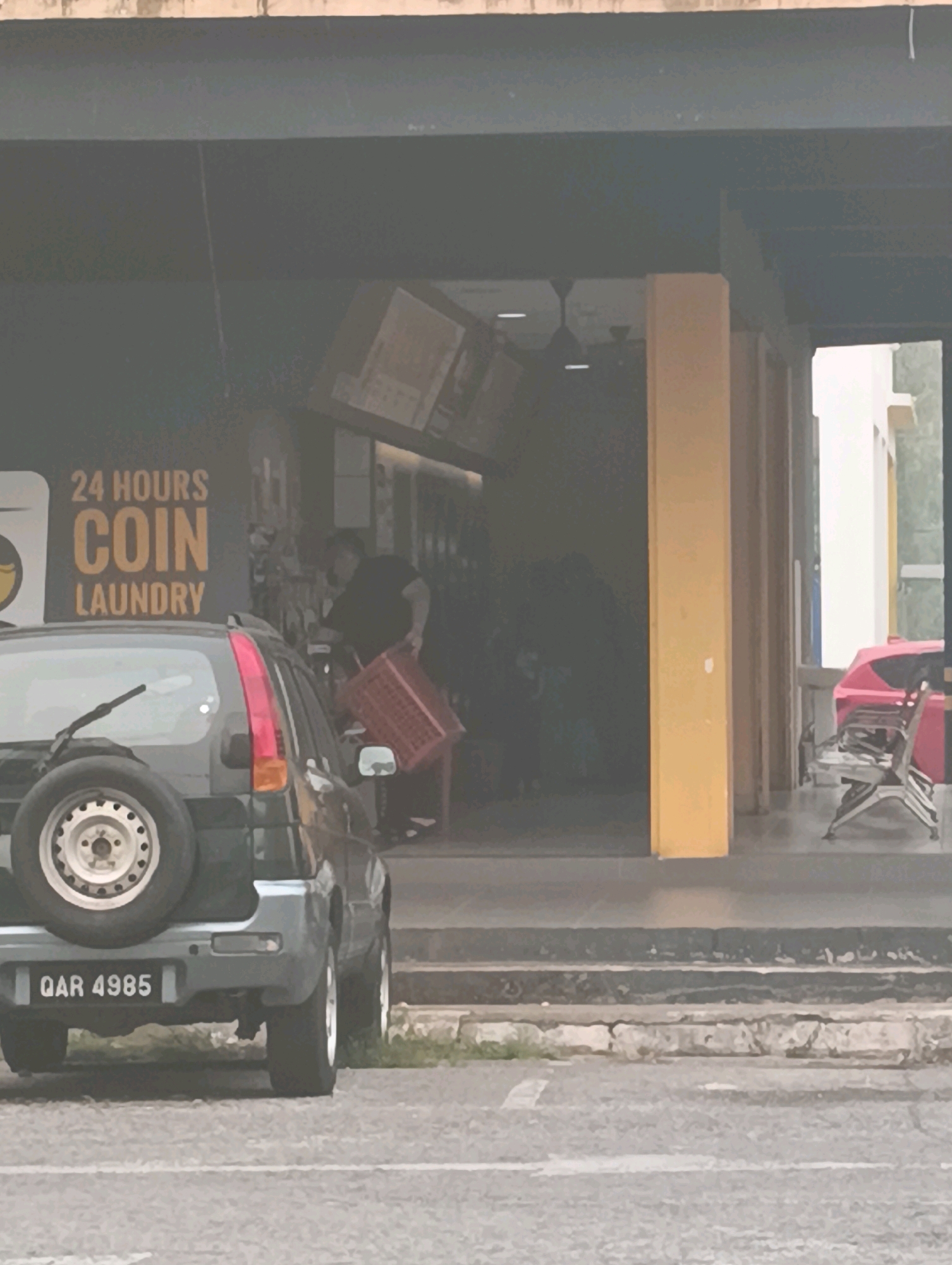 The image shows the exterior of a 24-hour coin laundry in Kuching, Malaysia. A dark-colored SUV with the license plate "QAR 4985" is parked in front of the establishment. Inside the laundry, a person wearing a black shirt and dark pants is visible, holding a red plastic basket. The building has a dark facade with orange lettering that reads "24 HOURS COIN LAUNDRY." A bright yellow pillar is present on the right side of the entrance. In the background, there are some chairs and a red object, possibly a trash bin, visible outside the laundry. The foreground consists of a paved parking area.