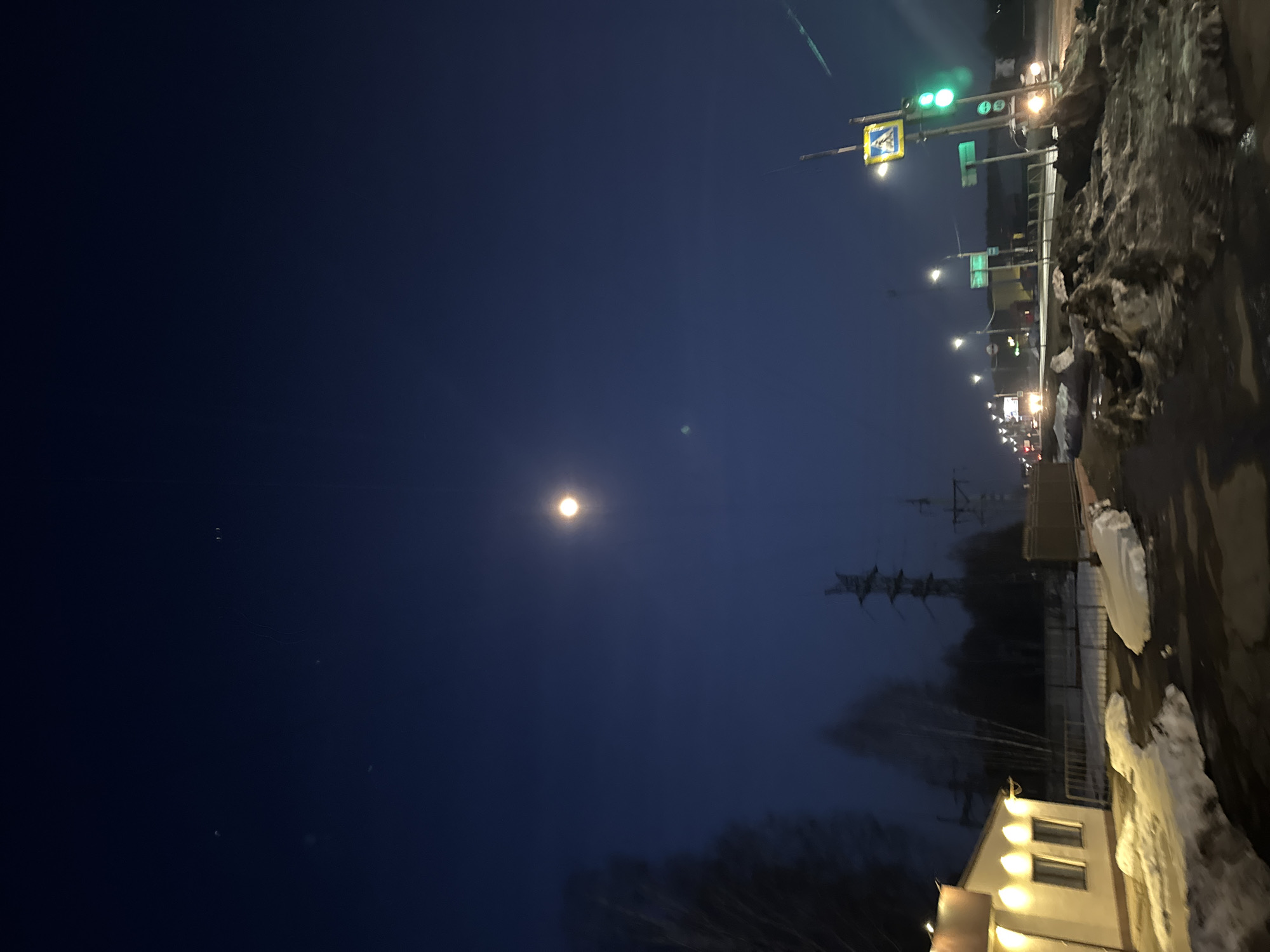 The image depicts a night scene in what appears to be Chaikovsky, Russia. The sky is dark blue with a visible moon. A street with traffic lights and streetlights illuminates the scene. There is a building on the bottom right with lights on. Snow and ice are visible on the side of the road. In the background, there are trees and what appears to be a power line.