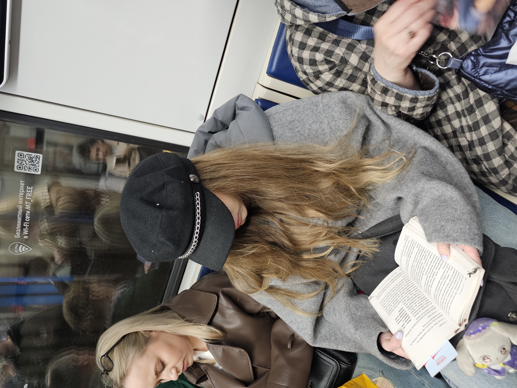 The image shows a group of people inside a train. A woman with long blonde hair is sitting down and reading a book. She is wearing a black hat with a silver chain, a grey coat, and blue jeans. Next to her sits another woman, wearing a brown jacket. The woman on the opposite side is wearing a checkered coat and holding a blue bag. There is a QR code and some Russian text on the window next to the first woman. The book has the word "ANSWER" visible on a bookmark. The train is likely in Khimki, Russia, due to the Russian text.