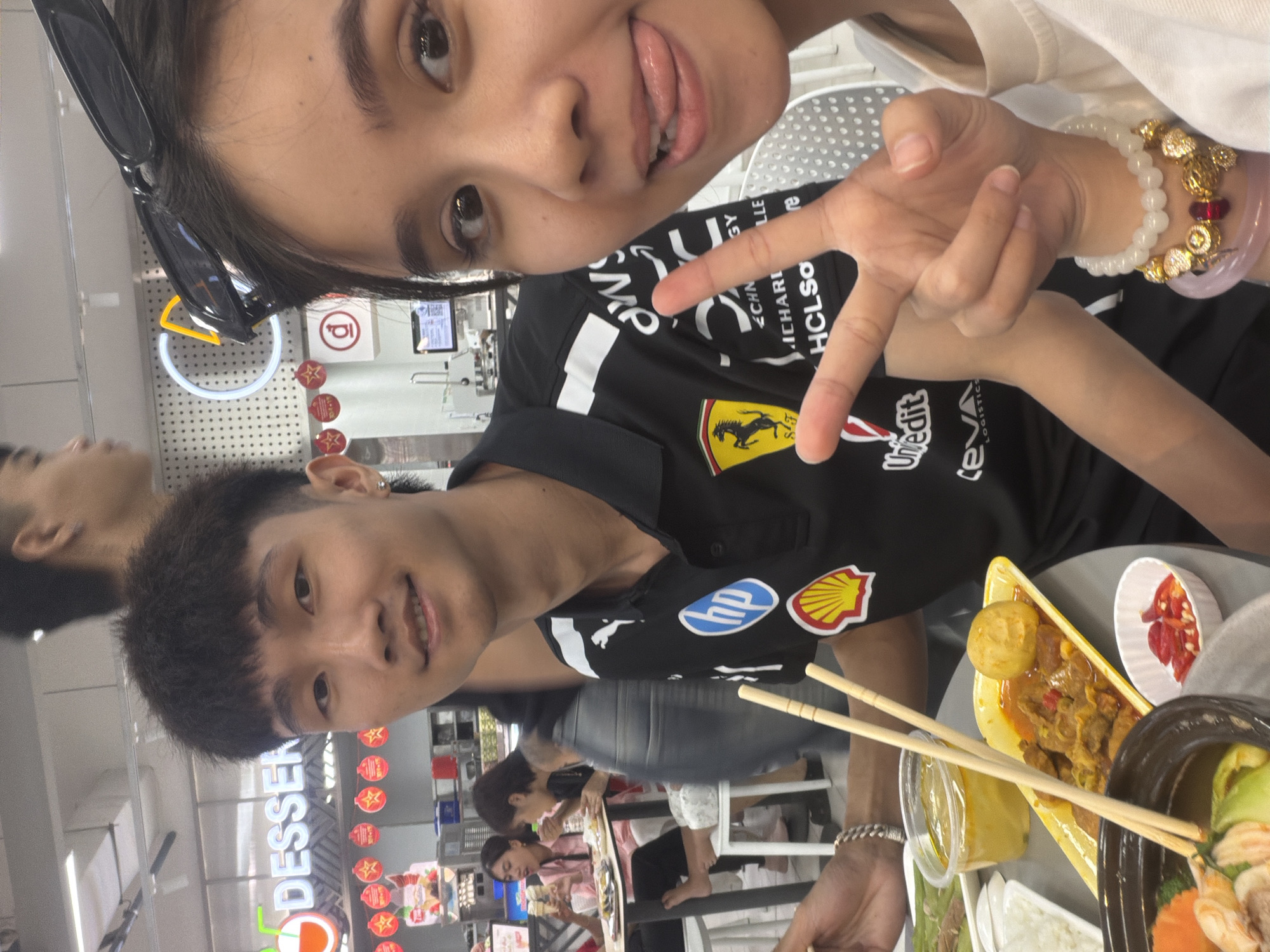 The image is a selfie taken indoors, possibly at a food court or market in Đà Nẵng, Vietnam. Two people are prominently featured. The person in the foreground is a younger man with short black hair, wearing a black shirt with various logos including Ferrari, Puma, HP, and Shell. He is looking at the camera with a slight smile. Behind him, another person, possibly a young woman, is also looking at the camera, sticking out her tongue and making a V-sign with her fingers. She has dark hair and is wearing a light-colored top. She is also wearing sunglasses on her head and a bracelet on her wrist.
In the foreground, food is visible: a yellow tray containing meat curry with a boiled egg, a small white bowl containing sliced chili peppers, and a black container with shrimp and vegetables. Chopsticks rest on the yellow tray.
In the background, other people are seated at tables, eating. A "DESSERT" sign with various decorations is visible on a pillar. A store display case with various packaged goods can also be seen in the background. The location seems to be a busy, commercial area with ample lighting.