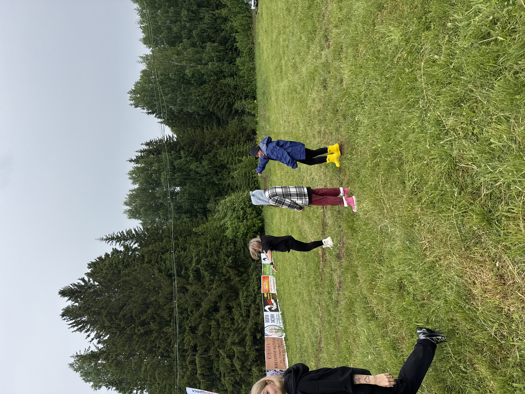 An outdoor scene captures several individuals in a grassy field with a dense forest line in the background under an overcast sky. In the foreground, partially visible, is a person wearing a black top, black pants, and black athletic shoes, with a tattoo visible on their left hand/wrist. Further into the midground, three people are standing. One wears a blue jacket, black pants, and bright yellow rain boots, appearing to bend forward. Another wears a plaid jacket, red pants, and pink shoes or boots. The third person in the midground wears a black top, black pants, and white shoes, bent over in a low stance, possibly stretching or exercising. The ground is covered in green grass, suggesting a natural, open environment. Along the edge of the forest, a series of banners or signs are visible. The visible text from these signs includes "БАНЩИКА", "ЮВИНА", "ЮВЕЛИРНАЯ МАСТЕРСКАЯ" (Jewelry Workshop), "ЛАСТОВКИ" (likely part of the workshop name), an address "ул. Степана Разина, 1" (Stepan Razin Street, 1), and a partial phone number "тел. 8 904 316". The setting is Bol'shaya Kivara, Russia, under what appears to be cloudy daytime weather, suitable for outdoor activity.