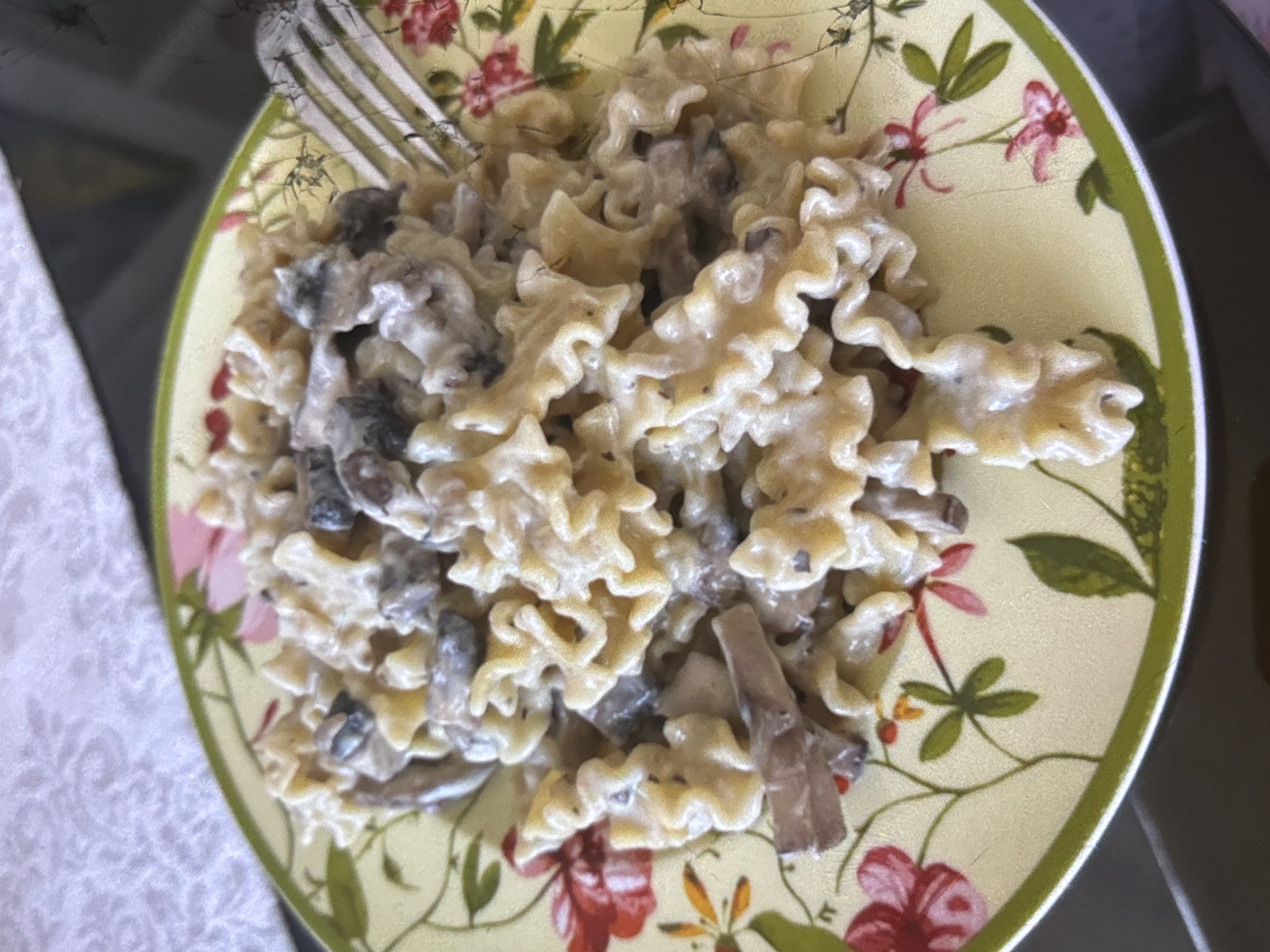 A plate of creamy mushroom pasta is presented on a decorative plate with a floral pattern. The plate is light yellow with green trim and features illustrations of pink and red flowers with green leaves and stems. A fork is partially visible on the right side of the plate, indicating the dish is ready to be eaten. The pasta appears to be ruffled or wavy in shape and is coated in a creamy sauce with visible mushroom pieces. The background shows a textured white surface, possibly a tablecloth, and a dark surface that might be a table. The scene is likely indoors. The image does not provide any information or visual cues to determine its location within the city of Orihuela, Spain.