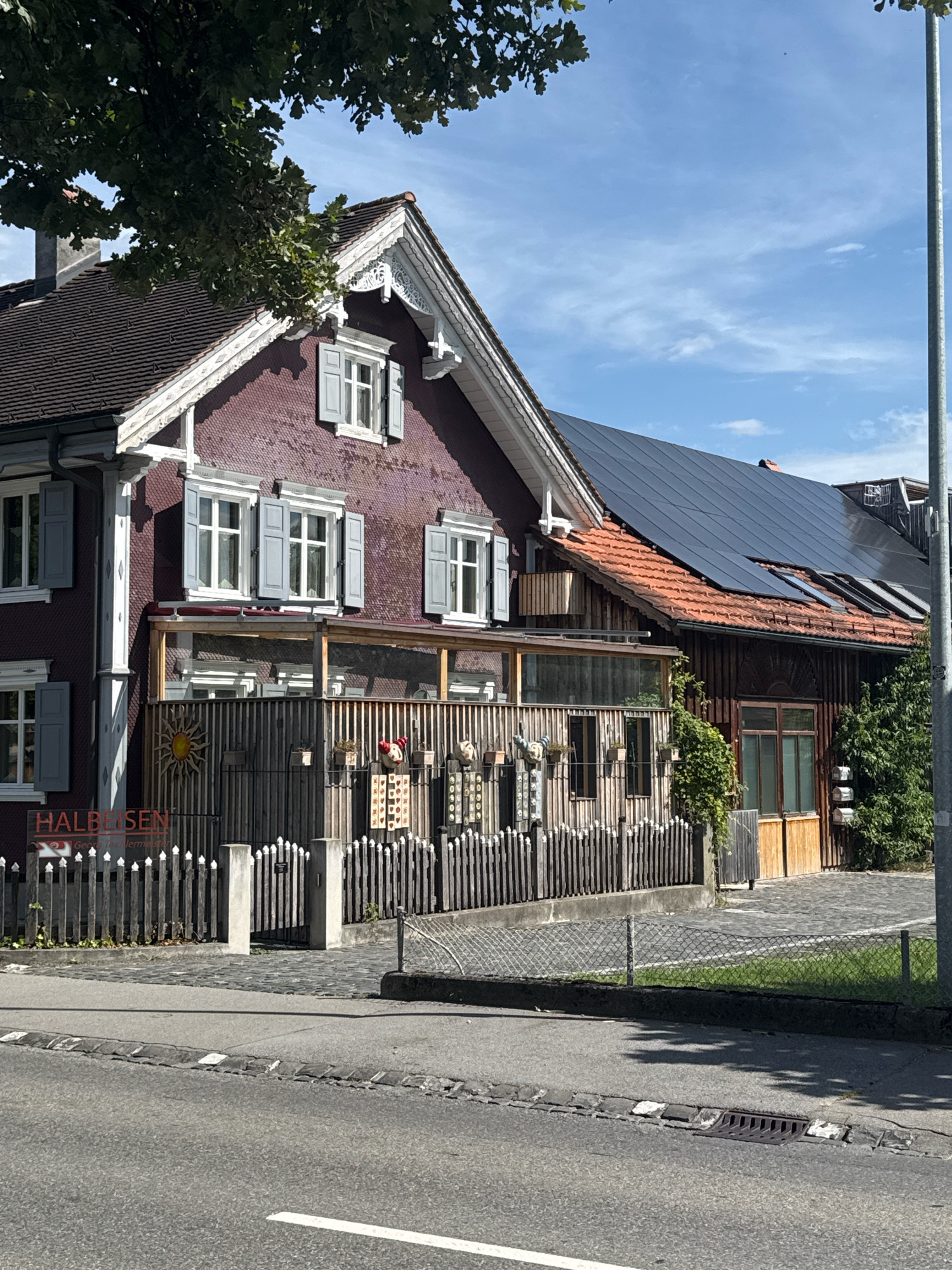An exterior, eye-level, medium shot captures a residential building with a dark red, textured exterior and a wooden extension. The main building features multiple windows with grey shutters and white trim. A section of the roof is covered in dark solar panels, while the other side has traditional orange tiles. A wooden fence runs along the front of the property, adorned with decorative elements like a sun and various figurines. Beyond the fence is a paved area and a street. The sky above is blue with scattered white clouds. A street light is visible on the right side of the frame, and a large tree with green leaves partially obscures the upper left corner. The scene appears to be in a suburban or rural setting.