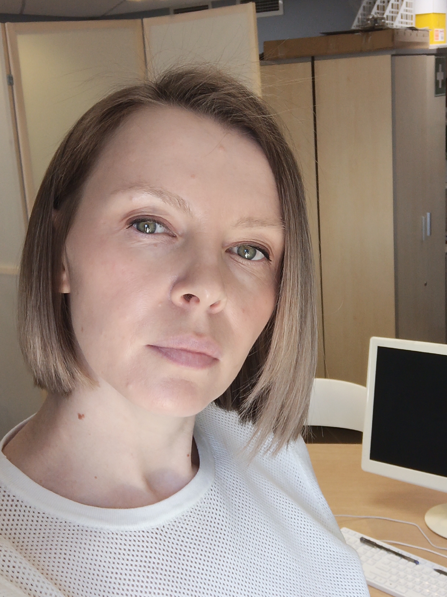 A close-up indoor shot depicts a Caucasian woman with fair skin, green eyes, and short, straight brown hair. She is wearing a white mesh-like top. Behind her are light-colored cabinets and a computer monitor. The background is indistinct but suggests an office environment. The city of Chaikovsky, Russia, cannot be determined from the image.
