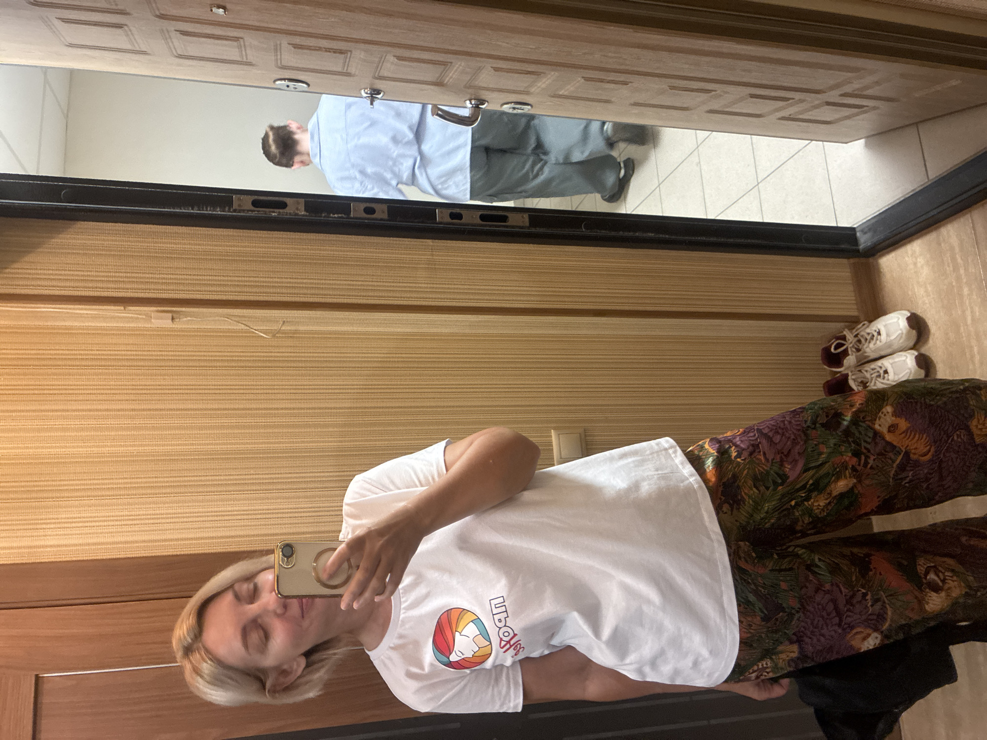 In a residential hallway in Sochi, Russia, a woman with blonde hair takes a selfie in a mirror. She is wearing a white t-shirt with a colorful circular logo and patterned pants. Her phone, held in her right hand, is visible as she captures the reflection. To her left, a black bag rests on the floor next to a pair of white sneakers. The hallway features a vertical textured wall and a wooden door. In the mirrored background, another person, seen from behind wearing a light blue shirt and gray pants, walks away through an open doorway. The floor is tiled in a light color.