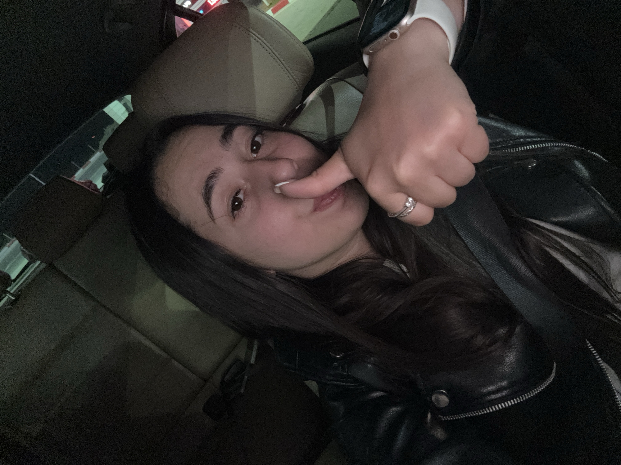 A selfie of a woman inside a car. She is wearing a black leather jacket and a watch on her left wrist, and a ring on her left ring finger. She is touching her nose with her left index finger. The car interior is visible, including a beige seat. The location in the city of Vodenichane, Bulgaria cannot be determined from the image.