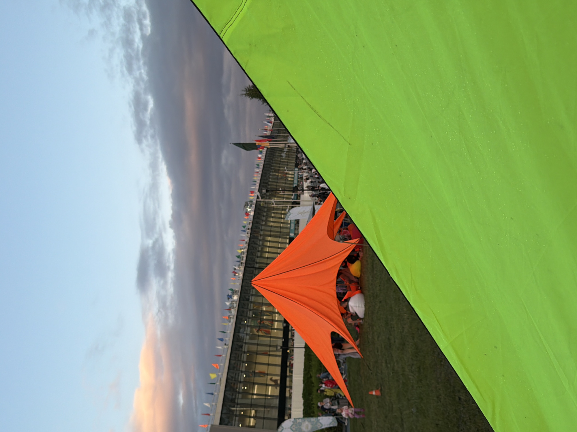 An outdoor scene shows a large, star-shaped orange tent in the center, with people sitting or standing underneath it. The tent is on a grassy field, and in the background, there is a long, modern building with glass windows. Flags of various colors are strung along the top of the building. To the right, a large portion of a bright lime green tent occupies the foreground, its surface showing small droplets of water. The sky above is a mix of blue and gray clouds, with hints of pink and orange suggesting it might be sunset or sunrise. Several people are visible on the grass around the orange tent and in front of the building. A small orange traffic cone is on the grass to the left of the orange tent.