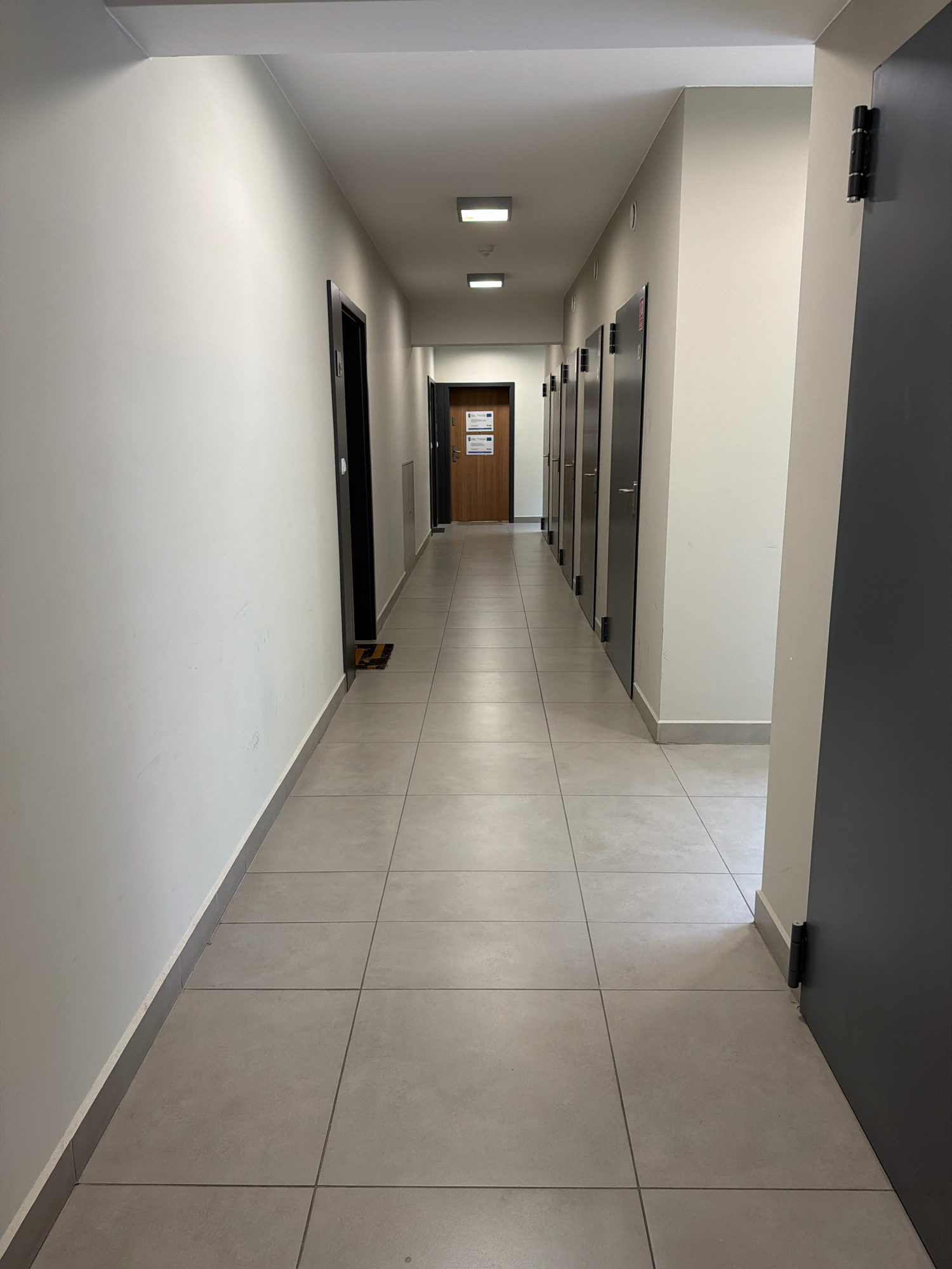 The image depicts a long, clean, and well-lit hallway in a modern building. The hallway is lined with multiple doors on the right side, all appearing to be dark gray or black with silver handles. The walls on the left are painted white, contrasting with the gray tiled floor. Rectangular light fixtures are recessed into the white ceiling. At the far end of the hallway, there is a wooden door with signage, suggesting an entrance or an office. A doormat with a yellow and black pattern is visible in front of one of the doors on the left, near the foreground. The overall impression is one of order and professionalism. Without further context, it is impossible to pinpoint the exact location in Wrocław, Poland.