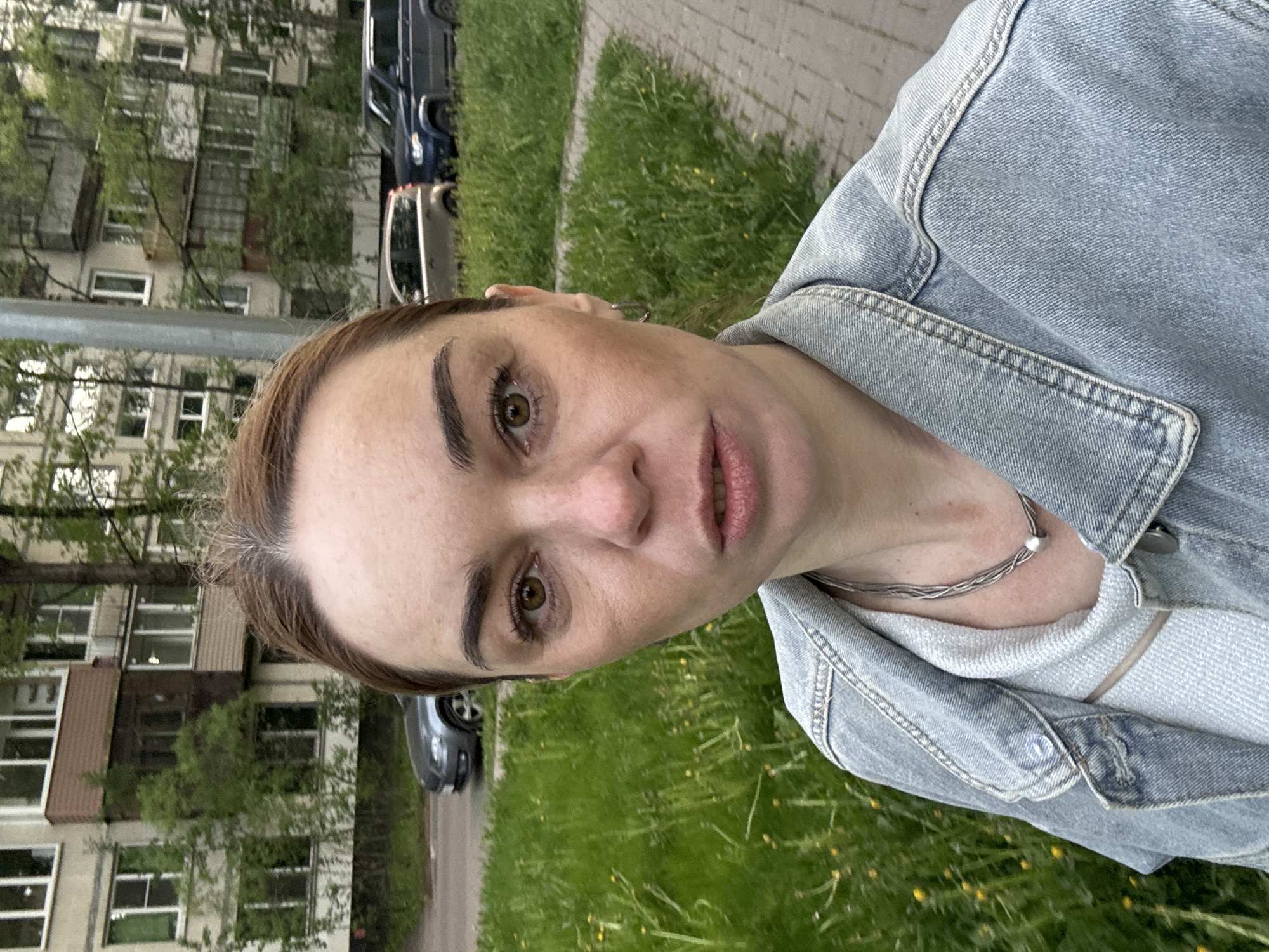 A woman is the primary subject in this medium shot. She is facing the camera, wearing a light blue denim jacket over a light-colored top and a silver necklace. The background shows a residential area, likely in Novoe Devyatkino, Russia. Buildings with multiple windows are visible on the left side, suggesting apartment blocks. Green grass and dandelions fill the space between the woman and the buildings. Vehicles, including a dark-colored van and a gray sedan, are parked along a road. The overall impression is an outdoor portrait against the backdrop of an urban landscape.