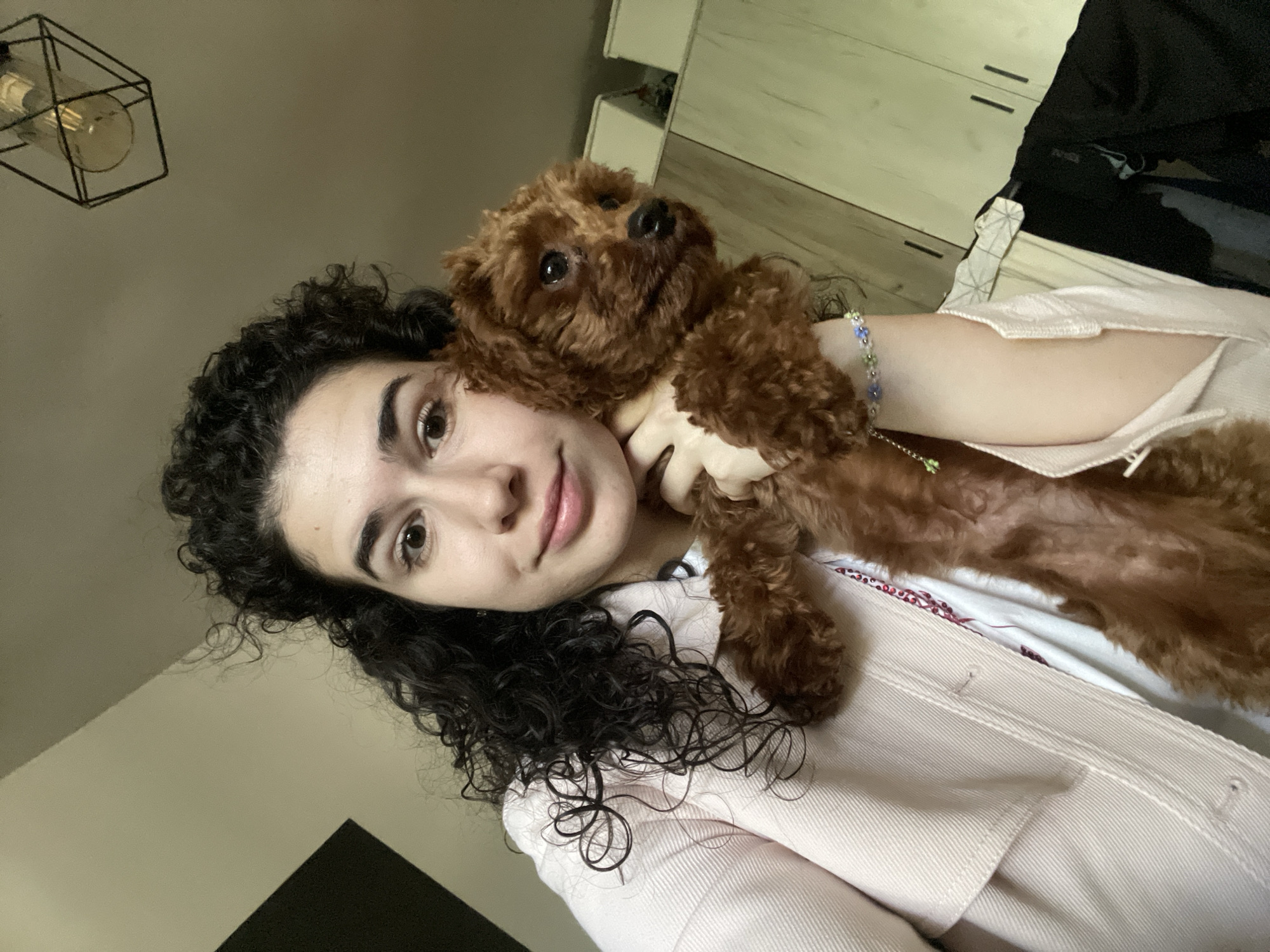 A selfie shows a woman holding a brown poodle. The woman has dark curly hair, fair skin, and is wearing a light pink jacket. The dog is held close to her face, and both are looking towards the camera. The background includes a light beige wall, a modern light fixture hanging from the ceiling, and a white cabinet with dark clothing inside. The location is presumed to be indoors but location specifics are not discernible.