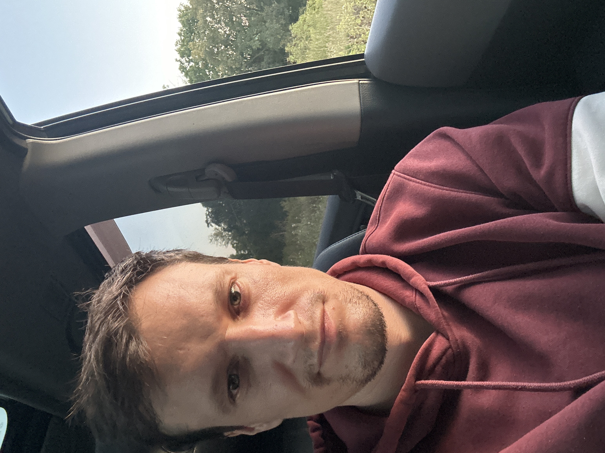 A medium shot captures a man from the chest up, seated inside a vehicle. He is looking directly at the camera, his expression neutral. The man has fair skin, short brown hair, and a light stubble on his chin and jawline. He is wearing a maroon hooded sweatshirt with drawstrings. The interior of the car is visible, with a black seatbelt mechanism on the right and the edge of a sunroof above. Through the car window on the right, a blurred background of green foliage and a bright sky suggests an outdoor, possibly rural, setting. The lighting indicates it is daytime, with sunlight casting shadows on the man's face.