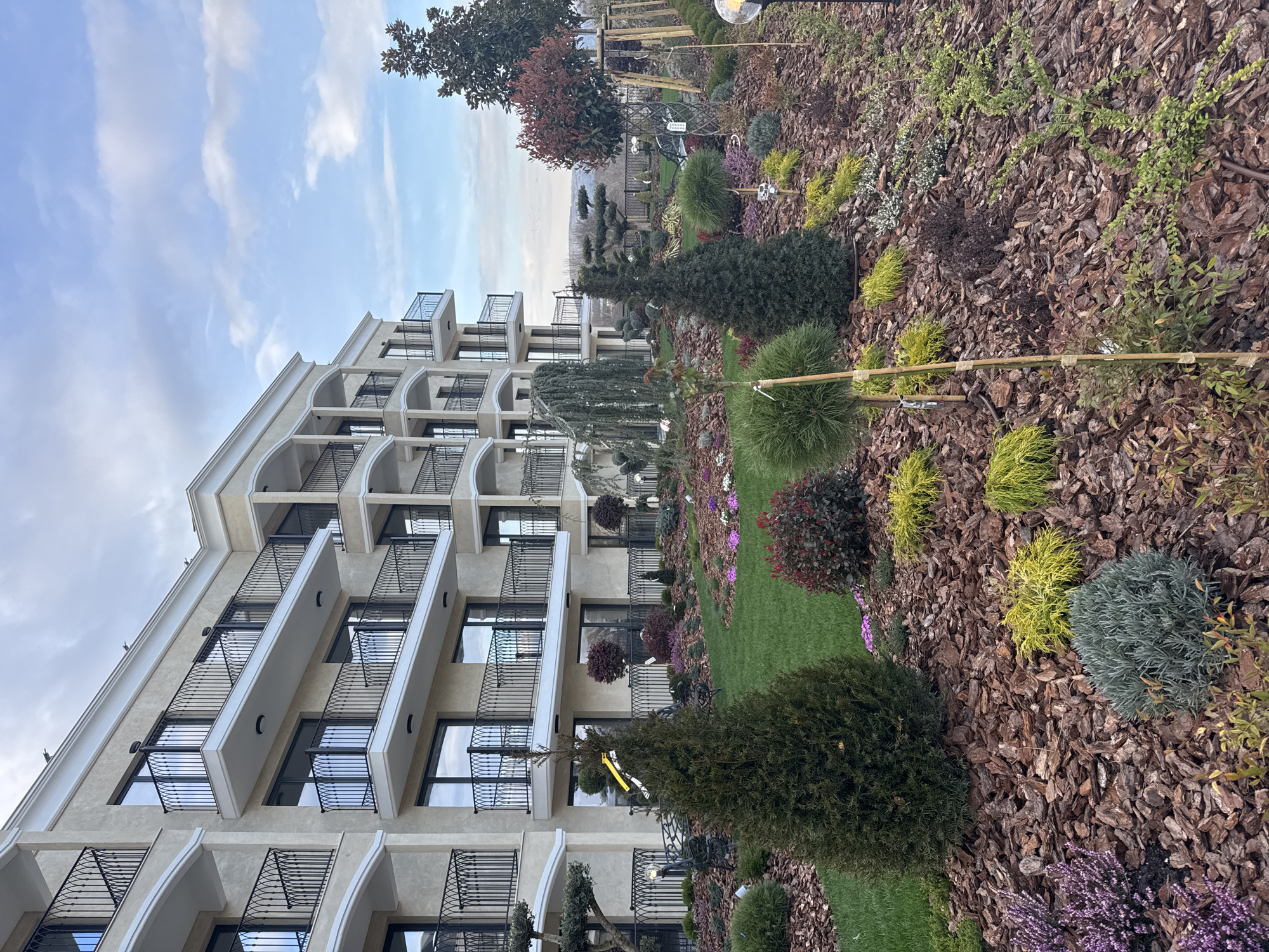 The image depicts a multi-story, light-colored building with numerous balconies and black metal railings. The building appears to be located in a well-maintained garden area featuring a variety of plants, including shrubs, trees, and flowering plants, and is partially covered with bark mulch. The sky is partly cloudy. Based on the prompt, the location is in the city of Kranevo, Bulgaria. No people are visible in the image.