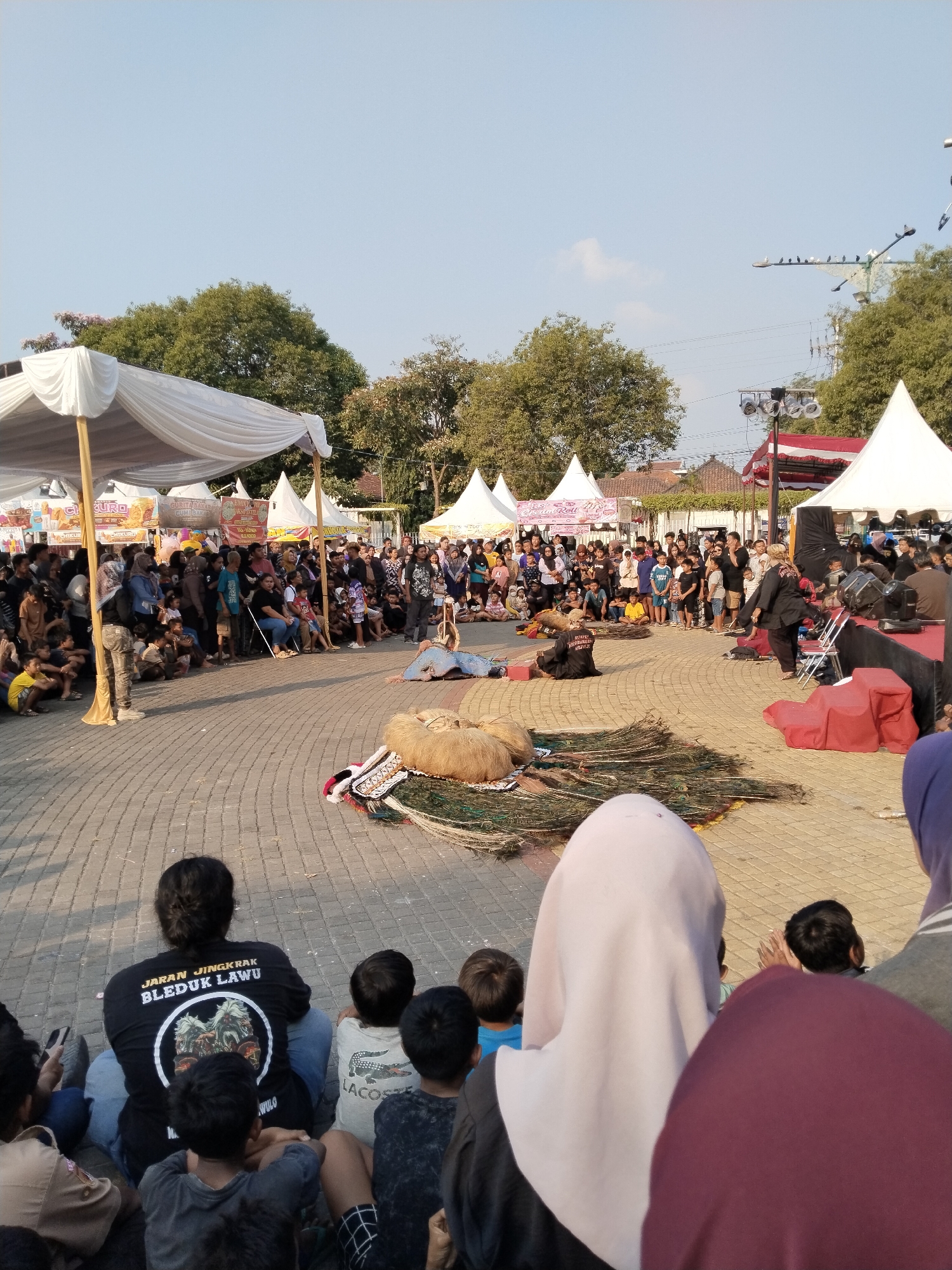 In Sragen, Indonesia, a street performance is taking place on a paved courtyard. A crowd of spectators, including children and adults, are seated on the ground watching the event. In the foreground, a person wearing a black t-shirt with the text "JARAN JINGKRAK BLEDUK LAWU" is seated. In the middle ground, several performers are on the ground, some dressed in elaborate costumes, with props such as peacock feathers and large, furry objects. Tents and stalls are set up in the background, indicating a festival or market atmosphere. The sky is clear and blue, and trees are visible in the distance.