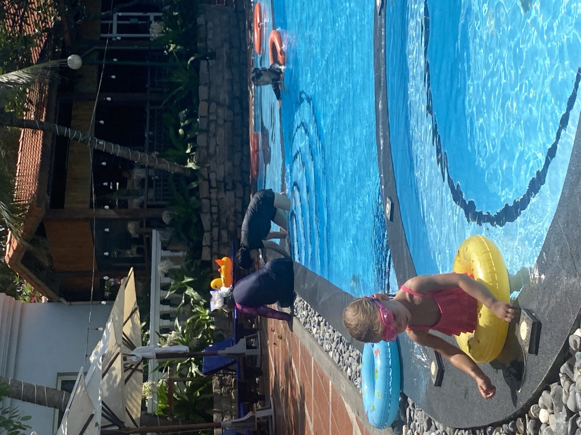 This image depicts a poolside scene in Mũi Né, Vietnam. In the foreground, a young girl wearing pink goggles and a pink ruffled swimsuit sits in a yellow inflatable ring in the shallow end of a bright blue swimming pool. She is reaching towards square objects embedded in the pool's edge. To her left, a light blue inflatable ring is partially submerged.
In the midground, two adults are partially visible in the deeper part of the pool. One adult, wearing a dark long-sleeved swimsuit, is crouched down. Another adult is swimming with an orange inflatable ring around their waist. Further back, another orange inflatable ring floats in the water.
The background reveals a resort or hotel setting. Umbrellas are set up poolside, and lounge chairs are visible. A building with a reddish-brown tiled roof and wooden accents is present. Lush green plants and palm trees surround the area, characteristic of Mũi Né's tropical environment. The lighting suggests it is daytime, with bright sunlight casting shadows.