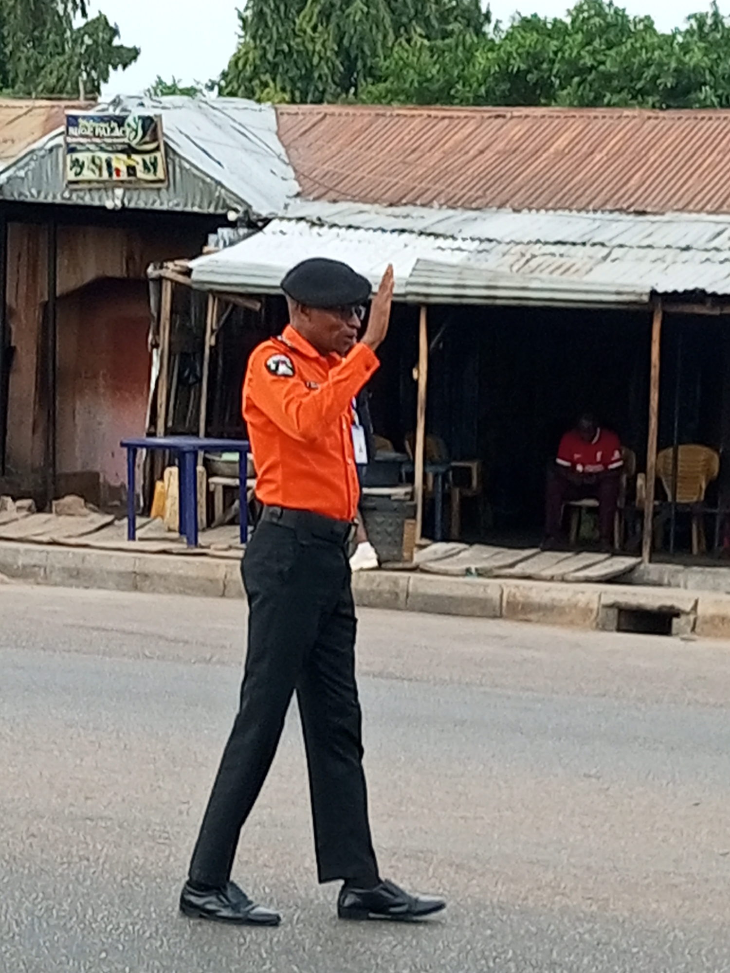 A person wearing an orange shirt and black pants, and a black beret, stands in a street in Tumfure, Nigeria, with their right hand raised in a greeting or stopping gesture. The person is positioned in the middle of the road, facing right with their body angled slightly towards the viewer. Behind them, a dilapidated building with a corrugated metal roof and wooden structures lines the street. A blue table is visible in front of the building, and a person in a red shirt is seated inside the building's darkened interior. The scene appears to be outdoors, with trees visible in the background.