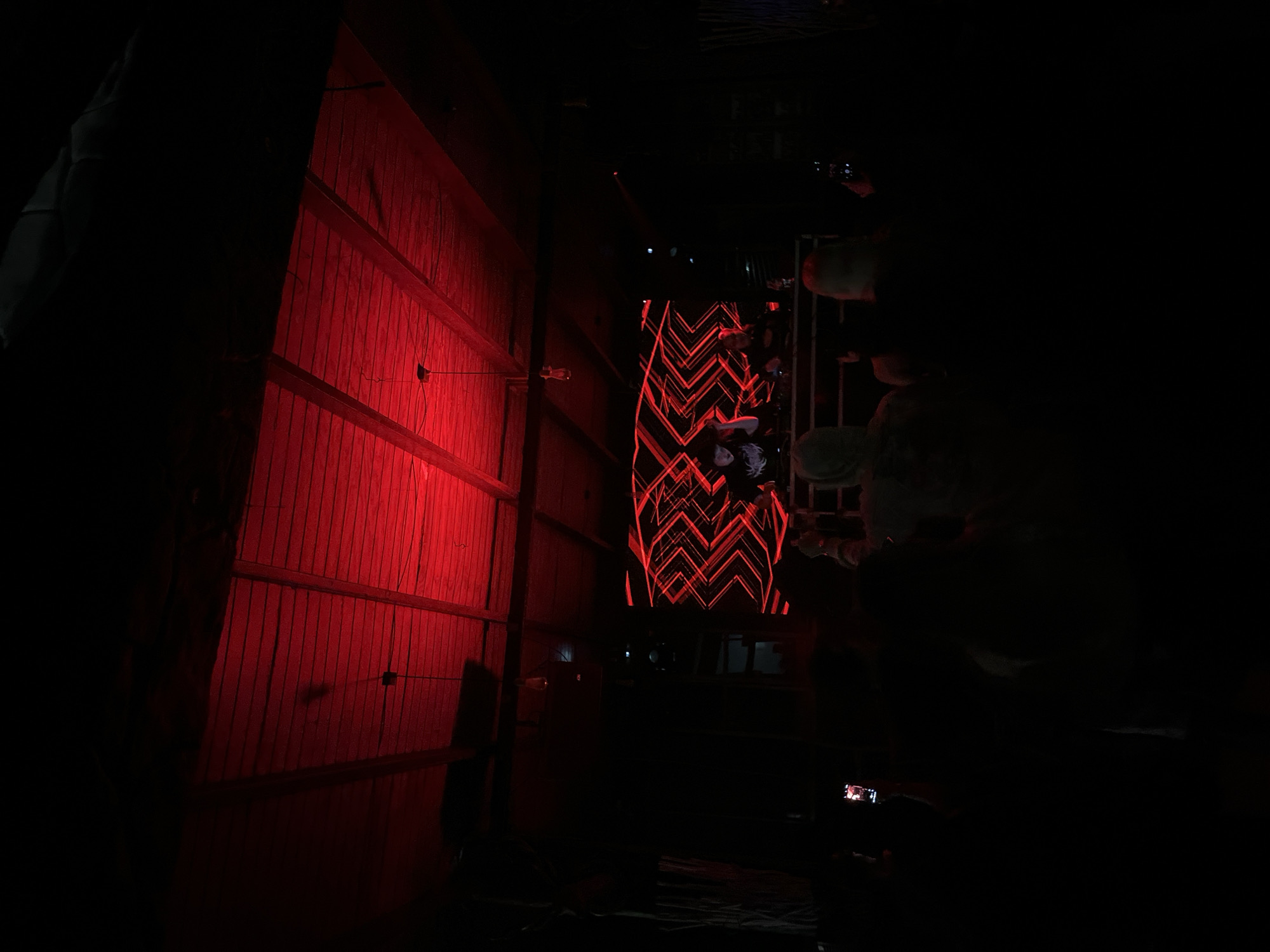 The image depicts an indoor scene, possibly a performance or event, in Arica, Chile. A wooden wall is illuminated with red light on the left side of the frame. A screen displaying a red geometric pattern is visible in the center, and figures of people can be seen in the background to the right of the screen. The rest of the scene is dark, with a person holding a phone in the foreground.