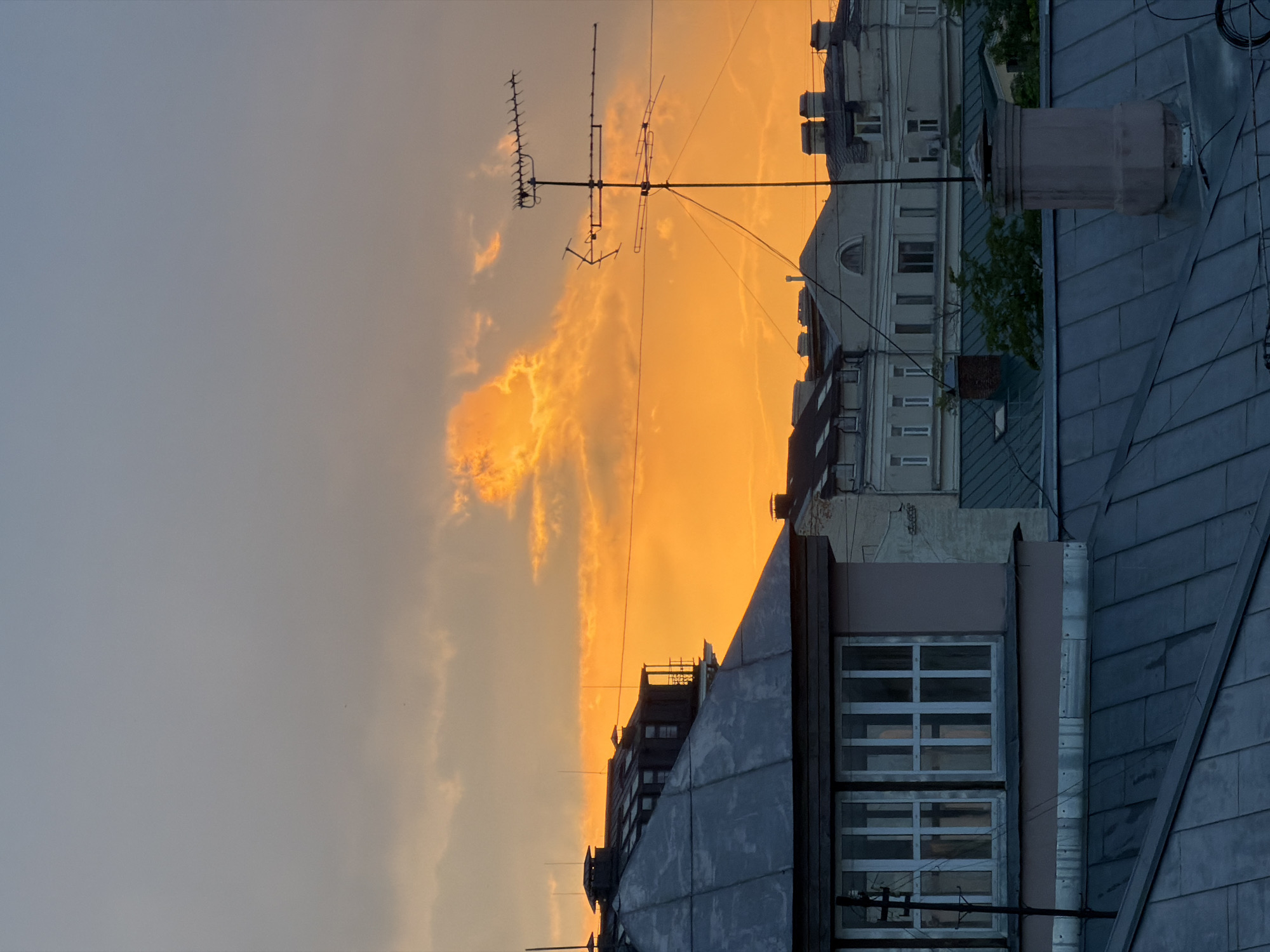 The image shows a cityscape at sunset, likely in Moscow, Russia. The foreground is dominated by rooftops covered in gray metal tiles, with visible windows and a chimney. In the background, multi-story buildings with various architectural styles are silhouetted against an orange and gray sky. An antenna is visible on a rooftop, extending into the sky. The overall environment suggests an urban setting with a mix of old and new buildings.