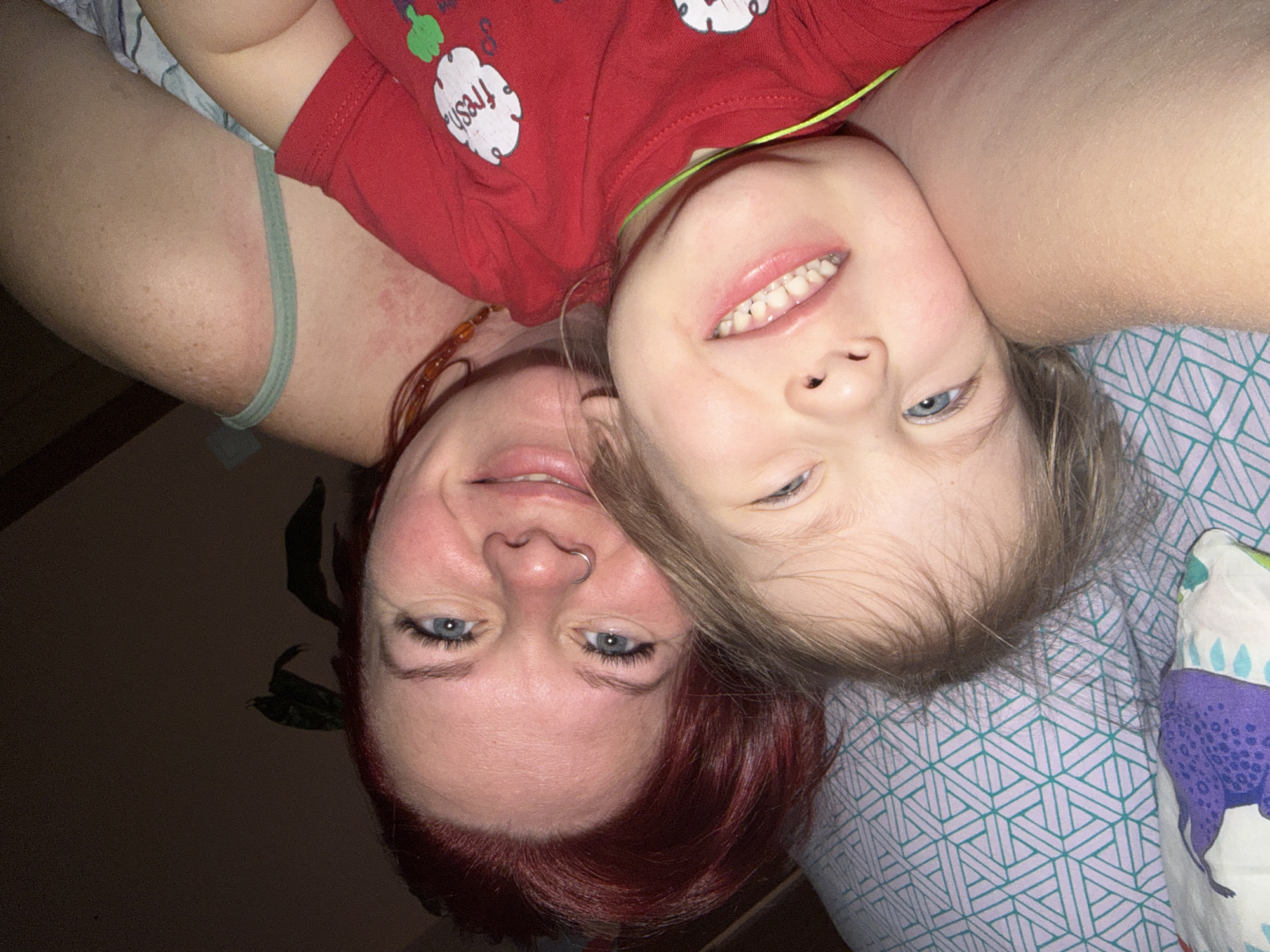A photograph depicts a fair-skinned woman with red hair and a nose ring lying upside down next to a young child wearing a red shirt with a "fresh" graphic. Both subjects have blue eyes and are smiling. The woman has a beaded necklace. The child's head rests on the woman's arm. The background includes bedding with a geometric pattern and a dark, shadowy area, possibly a wall with a plant. The photo's location is not discernible from the image.