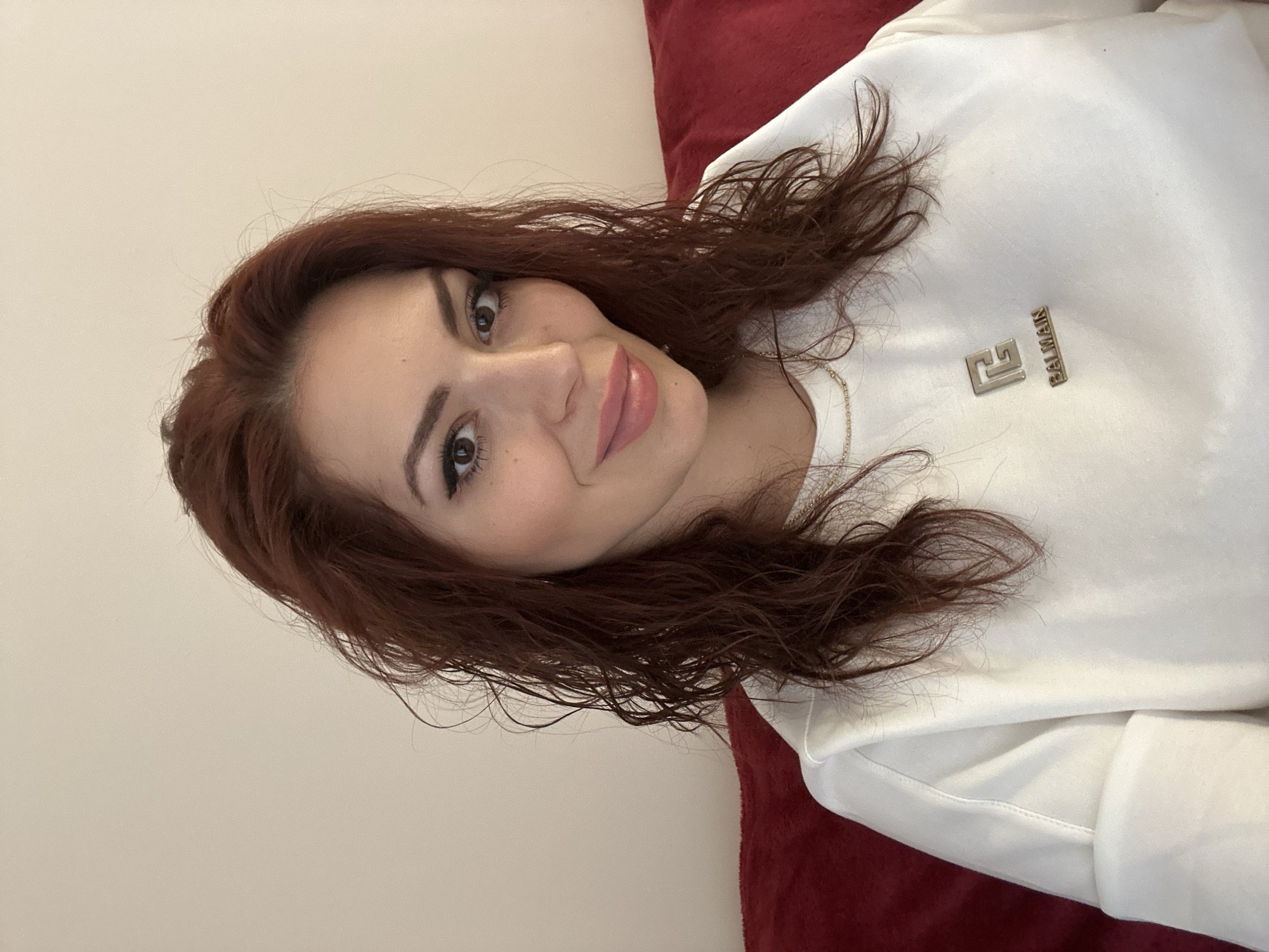 A female subject with reddish-brown, wavy hair, and brown eyes is depicted in a close-up, chest-up shot. She has visible makeup, including eyeliner and light pink lip gloss, and is smiling subtly while looking directly at the camera. She is wearing a long-sleeved white sweatshirt or sweater with cuffed sleeves. A metallic gold-colored emblem displaying a stylized "G" or "B" over the word "BALMAIN" in capital letters is affixed to the front center of her top. A thin, gold-colored chain necklace is visible around her neck. The background consists of a plain, light beige wall to her left and a portion of a red upholstered surface, likely a sofa, to her right and beneath her. The lighting is soft and even. This indoor scene is situated in the city of Sofia, Bulgaria.
