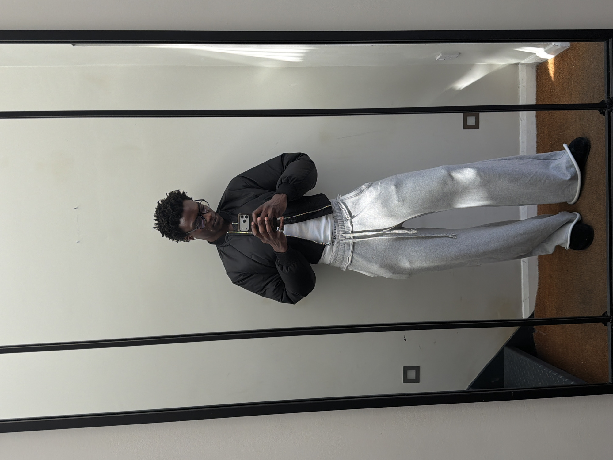 The image depicts a man reflected in a full-length mirror. He is wearing glasses, a black bomber jacket over a white shirt, and grey sweatpants with black shoes. He is holding a smartphone, presumably taking the photo. The mirror has a black frame and reflects a room with light-colored walls, a corkboard floor in the top right corner and two electrical outlets visible on the wall. Given the architectural style and general aesthetic, the location may be in London, United Kingdom.