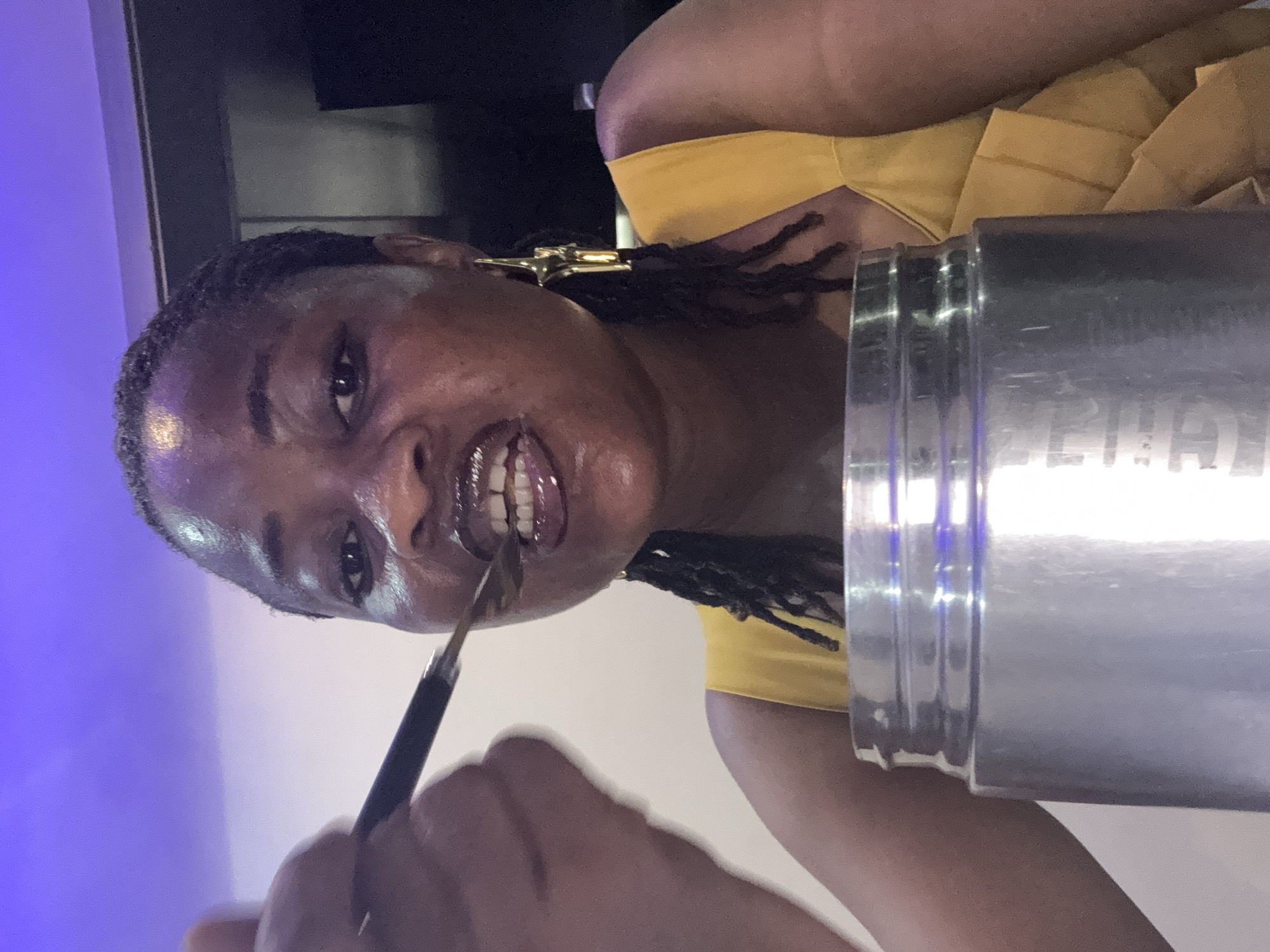 A medium shot depicts a Black woman with dark, braided hair adorned with gold cross earrings. She is wearing a yellow top and holding a black-handled makeup brush to her lips, apparently applying lip gloss. A tall, silver-colored, cylindrical container with indiscernible writing on its surface is prominently positioned near her. The background shows a wall with a dual color scheme, featuring a purple area on the left and a white section on the right. There is no information available to confirm the location within the city of Ajara, Nigeria, or any context about the environment or other interactions.