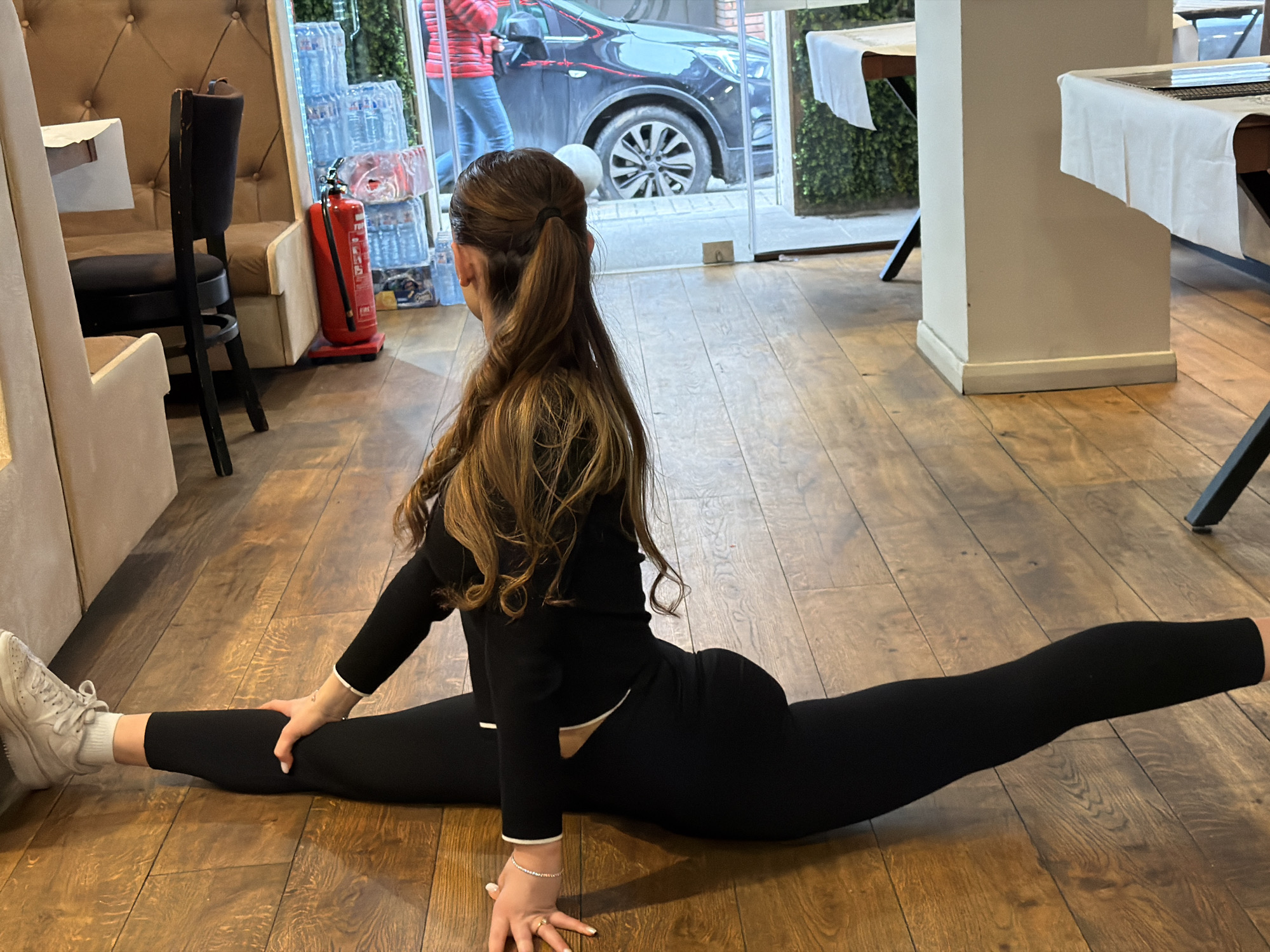 A woman with long, wavy brown hair pulled into a ponytail is performing a split on a wooden floor inside a building. She is wearing a black long-sleeved top and black leggings, and white sneakers. She is facing away from the camera.
Behind her is a glass window with a partial view of a black SUV parked on the street. In the background to the left, there is a beige padded booth seat with a black chair. A red fire extinguisher is standing near the window. To the right of the woman, tables with white tablecloths are visible.
Based on the style of furniture and ambiance, the location appears to be a restaurant or cafe. Considering the license plate on the vehicle outside, it is highly probable that the location is in Sofia, Bulgaria.