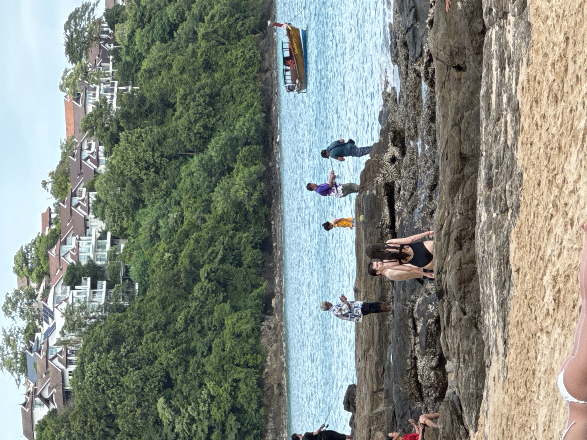 The image depicts a coastal scene in Wichit, Thailand, featuring a rocky shoreline in the foreground and a body of water beyond. In the background, a verdant hillside is densely covered with trees, atop which are several multi-story buildings with white walls and terracotta roofs. The water is a bright blue, and a traditional wooden boat with a canopy is visible on the sea.
Several people are present. On the rocky shore, a man wearing a black shirt and pants is standing and holding a fishing rod. Nearby, an older man in a patterned shirt and black shorts is looking at his phone. Another man in a green shirt and camouflage pants is also present. Further along the shore, a young child in an orange garment is standing. In the mid-ground, on the rocks, a couple is visible; the man is shirtless, and the woman is wearing a black swimsuit. Closer to the foreground, on the sand, a person wearing a white bikini bottom is lying down, facing away from the camera.
The environment includes textured rocks, clear blue water, and lush green foliage. The sky is overcast, and the overall lighting suggests daytime.