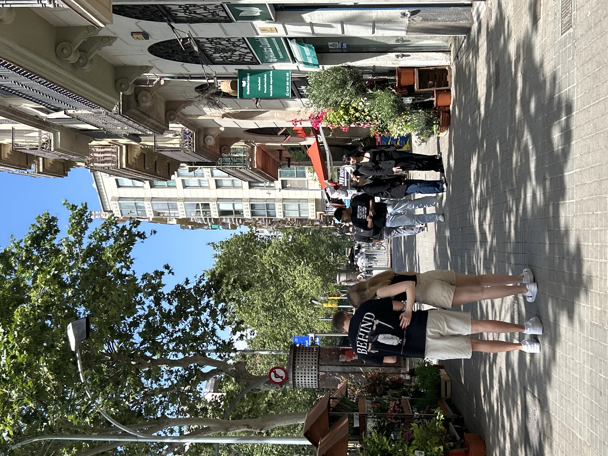 The image captures a street scene in Barcelona, Spain, characterized by a paved walkway and a row of buildings on the left side. People are seen walking along the pavement, some in groups and others in pairs. The buildings feature architectural details such as ornate balconies and awnings. A dense tree canopy partially obscures the sky. Street signs and storefronts are visible, with vegetation adding a touch of greenery to the urban environment. The overall scene conveys a sense of everyday life in Barcelona.