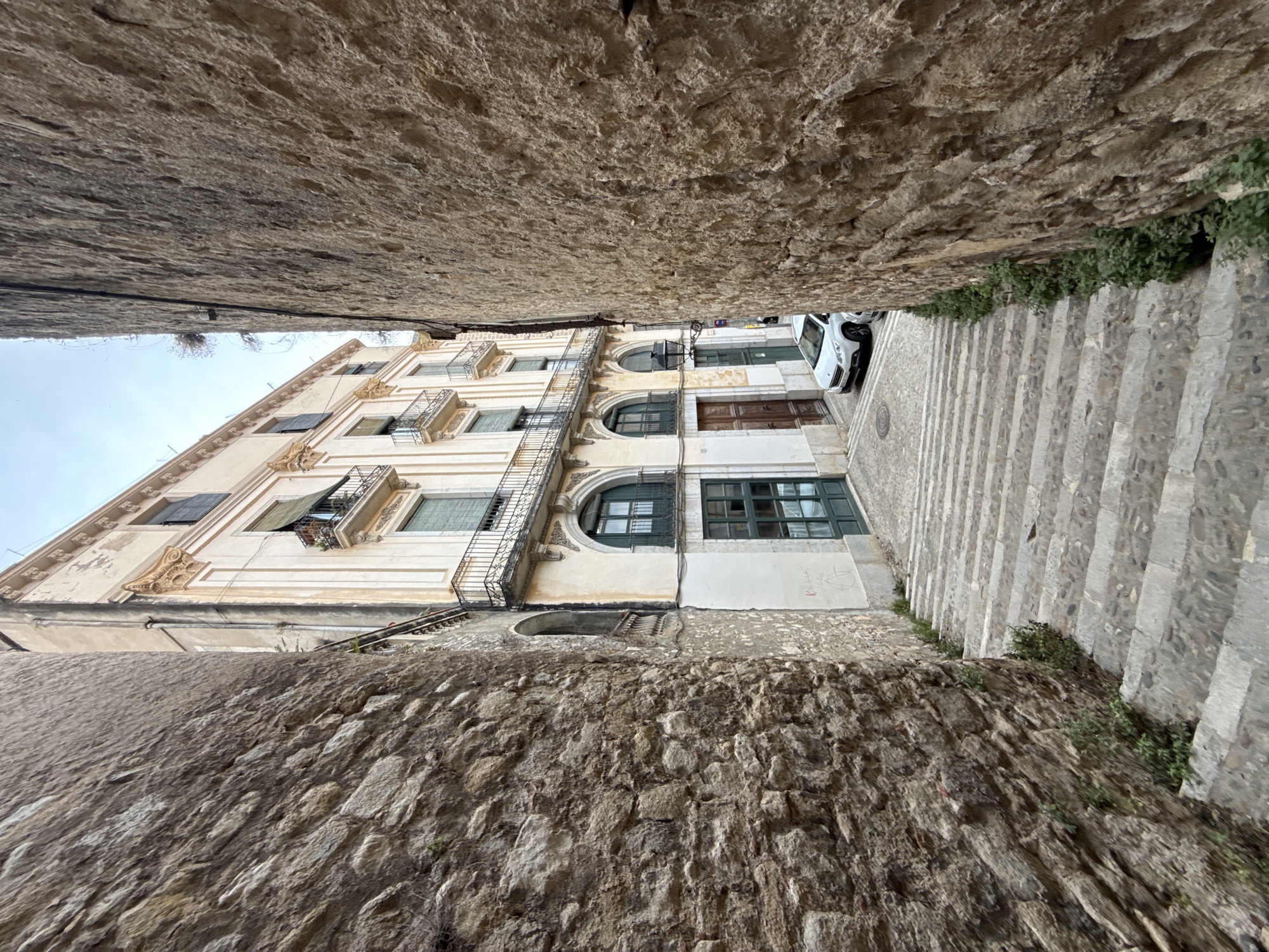 The image shows a narrow, steep stone staircase leading downwards between two old stone walls. On the right, the wall is rough-hewn and textured. On the left, the wall is also stone but appears to be built with larger, more irregular stones. At the bottom of the stairs, a white car is parked on a paved street. Beyond the car, a building with ornate architectural details, including balconies and arched windows with green shutters, rises up. The building is painted a light cream color and has visible signs of age, with some peeling paint. The sky above is overcast. The scene is located in the historic center of Girona, Spain, likely in a Gothic Quarter alleyway. There are no people visible in the image.