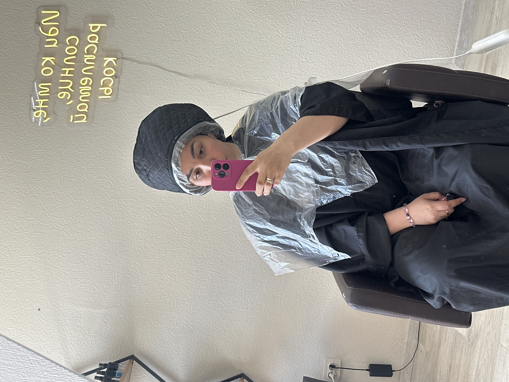 The image captures a woman sitting in a salon chair with a black quilted cap on her head. She is wearing a black salon gown and a clear plastic cape, and holding a pink smartphone in her right hand, as if taking a selfie. The background features a light beige wall with a neon sign written in an unidentified language. A white electrical cord dangles from the wall towards the ceiling. The floor has light-colored wooden planks. Other items such as a shelf, and the power adapter are visible in the background. The location appears to be a salon in Unknown, Unknown.