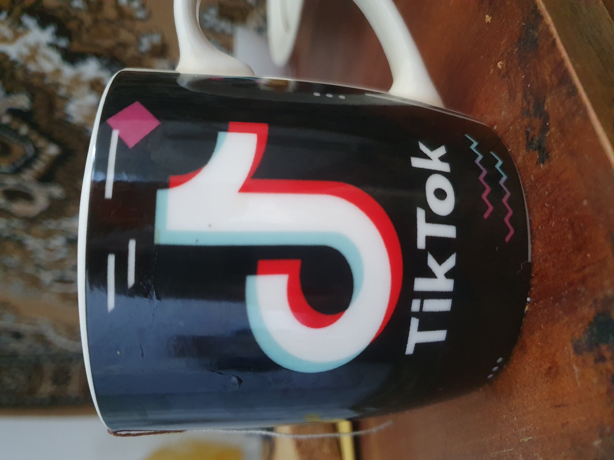 A close-up, eye-level shot captures a black mug featuring the TikTok logo and text. The logo is a stylized white "J" with red and cyan outlines, and the text "TikTok" is in white below it. Two wavy lines, one pink and one blue, are positioned beneath the text. The mug has a white handle and a white rim. The background is blurred, showing a patterned fabric tapestry and a wooden surface, possibly a table. A small portion of a tea bag string is visible hanging over the mug's left side. The scene is set indoors, on what appears to be a wooden table, and there are no people visible in the image. The location of Zubrești, Moldova is not discernible from the visual information provided in the image.